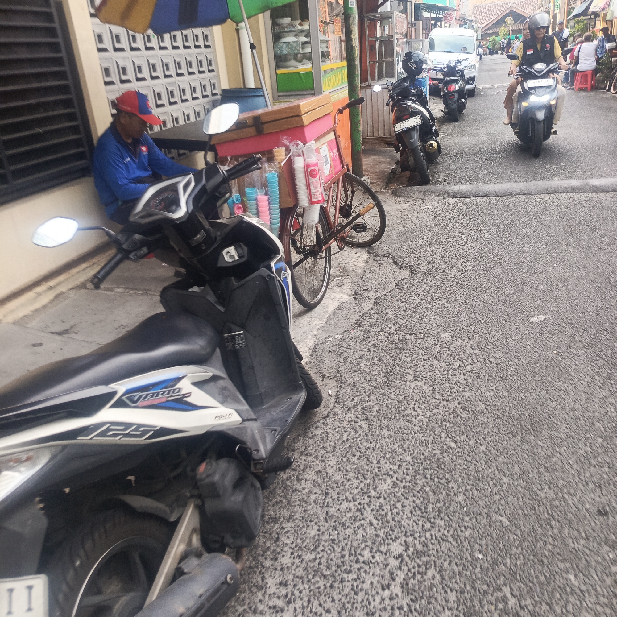 A street scene in South Jakarta, Indonesia, captures a moment of daily life. In the foreground, a white and black scooter is parked on a grey asphalt road. To the left, a man wearing a red and blue cap and a blue jacket sits beside the scooter, facing a table with an umbrella overhead. Behind him, a cart with various colorful plastic cups and containers is attached to a bicycle. Further down the road, several motorcycles are parked or in motion. One motorcycle, a black one with a license plate reading "T 2370 MA," is visible. Another black motorcycle is ridden by two people, a woman in front and a man behind wearing a helmet and a yellow vest. In the background, buildings and shops line the street, and a white van is also present. The overall impression is a bustling urban environment, typical of Indonesia.