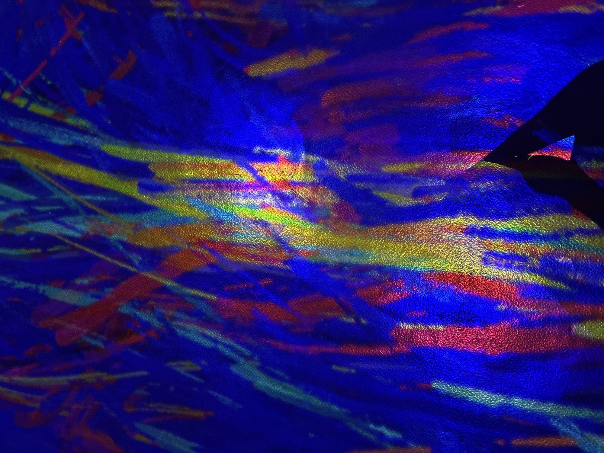 The image is an abstract and colorful photograph, likely taken in Abu Dhabi, United Arab Emirates. The main subject appears to be a textured surface, possibly a wall or fabric, illuminated by projected light that creates a vibrant, abstract pattern. Streaks and splashes of various colors, including deep blue, red, yellow, and green, are visible, creating a dynamic and energetic visual effect. A dark, silhouetted object, triangular in shape, is present in the lower right portion of the frame, partially obscuring the colored pattern. This silhouette could be a piece of furniture, a sculpture, or a shadow cast by an unseen object. The overall impression is one of artistic expression and visual exploration, with the play of light and shadow on the textured surface being the primary focus. There is no specific identifiable landmark of Abu Dhabi visible.