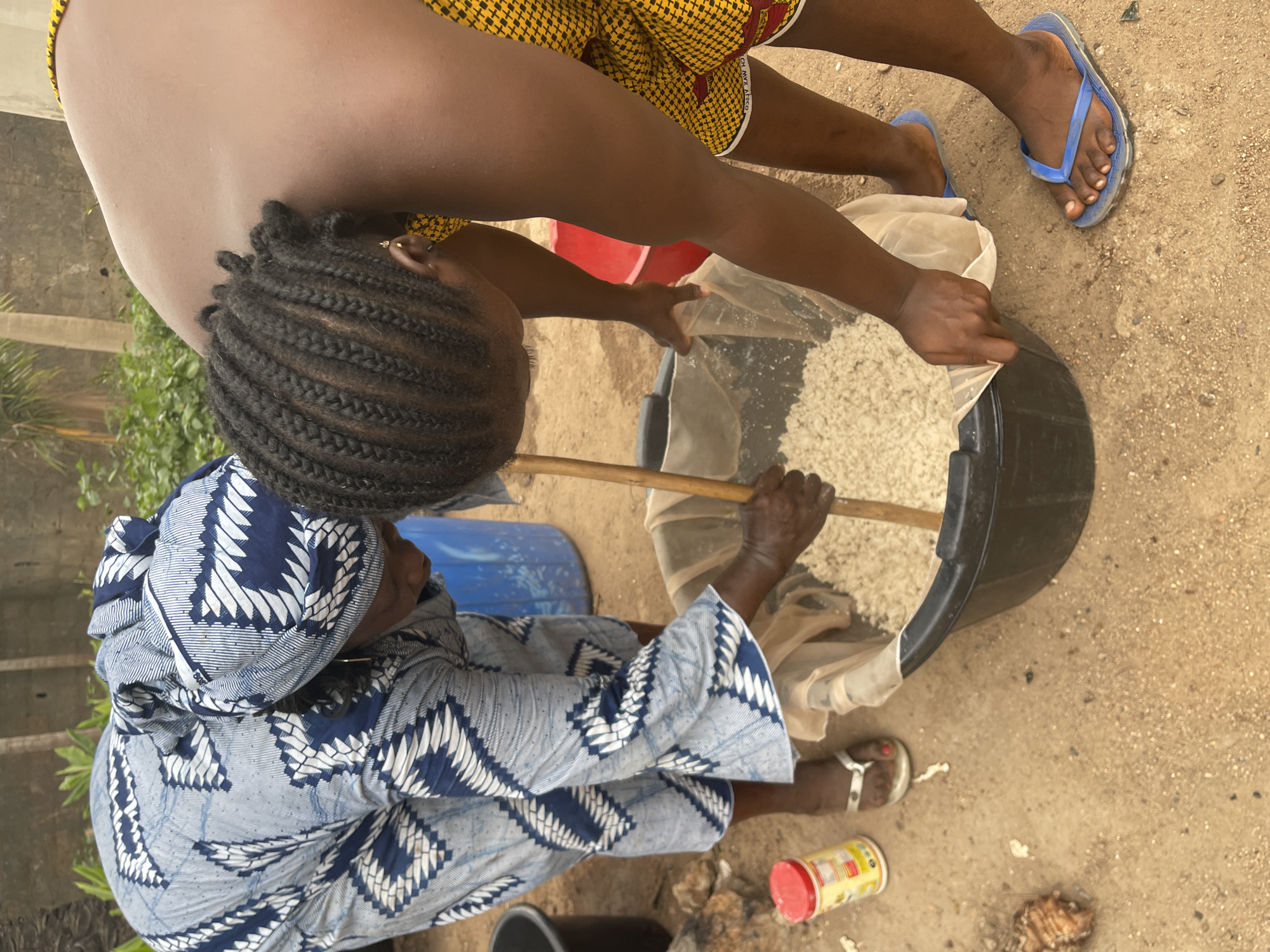 A high-angle shot shows two individuals engaged in a task, likely related to food preparation or processing, outdoors in Akwanga, Nigeria. The ground is sandy and scattered with small debris, including what appears to be a discarded container.
One person, dressed in a blue and white patterned headwrap and matching top, is crouching and holding a wooden stick or tool inside a large, black plastic tub. The tub is lined with a white, porous cloth, possibly a filter, containing a mass of light-colored material, resembling rice or another grain.
The second person, with braided hair and wearing yellow patterned shorts, leans over the tub, also holding the edge of the filter or cloth. The person is assisting in the process or observing the work being done.
The environment suggests a rural or semi-rural setting, potentially within a residential area.