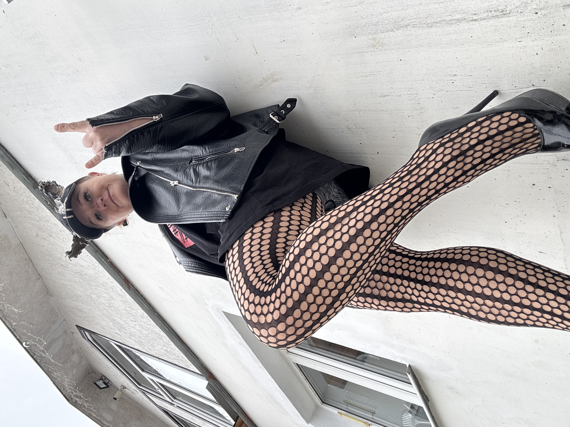 A young woman stands in Chaikovsky, Russia, angled slightly upwards towards the camera. She wears black fishnet stockings, high-heeled black patent leather shoes, a black mini skirt, a black t-shirt, and a black leather jacket. She also wears a black cap. Her left hand is raised, making a "rock on" gesture. The background features a building with white walls, windows, and a surveillance camera. The sky appears overcast.