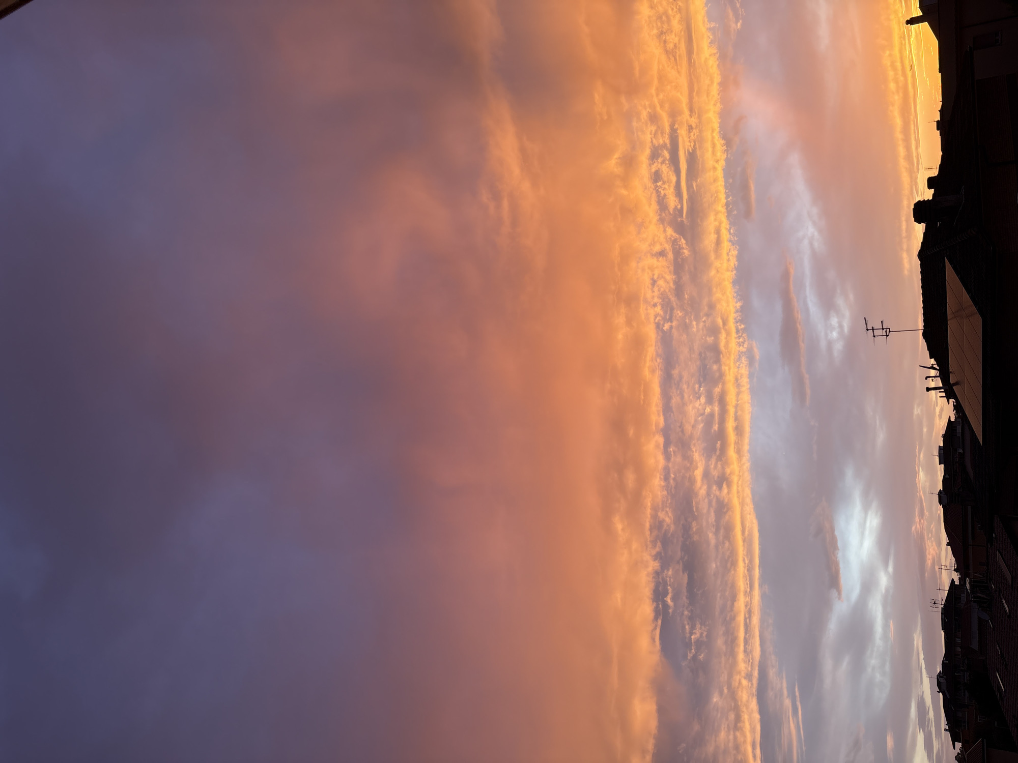 The image shows a colorful sky at sunset, with shades of orange, pink, and purple. The sky is filled with clouds, some of which are illuminated by the setting sun. In the lower right corner of the image, there are silhouettes of buildings. The location is a city; the city name and region are Unknown.