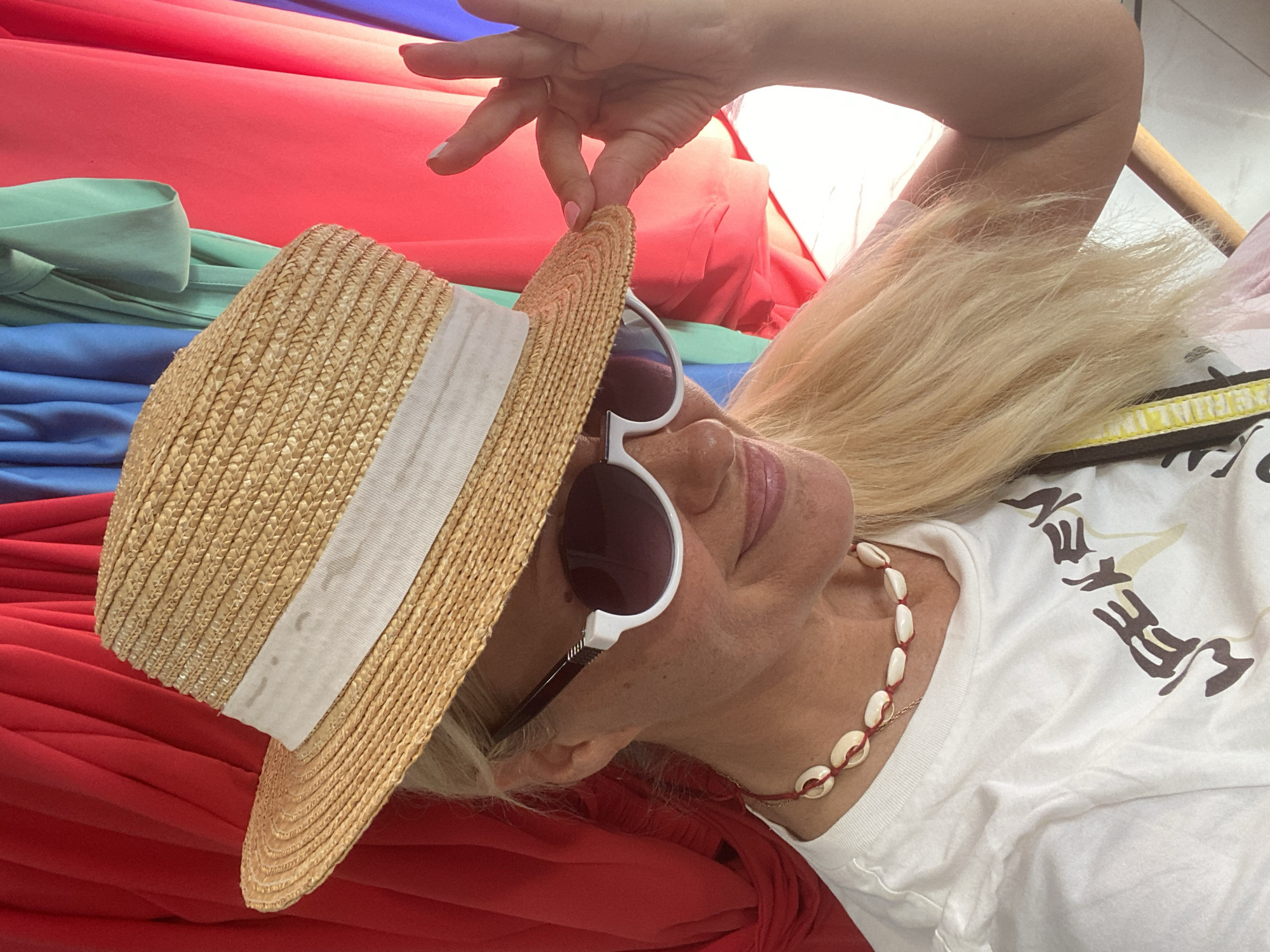 A blonde woman wearing a straw hat, white sunglasses, and a white t-shirt with the word "WEEKEND" printed on it stands in front of a colorful background of draped fabric. She is also wearing a necklace made of shells. The woman is looking down and to the side, and her right hand is raised, touching the brim of her hat. The background appears to be indoors, possibly a boutique or a dressing room, with red, blue, and green fabric visible. The lighting is bright, suggesting it might be daytime. The specific location in Thira Municipal Unit, Greece cannot be determined from the image alone.