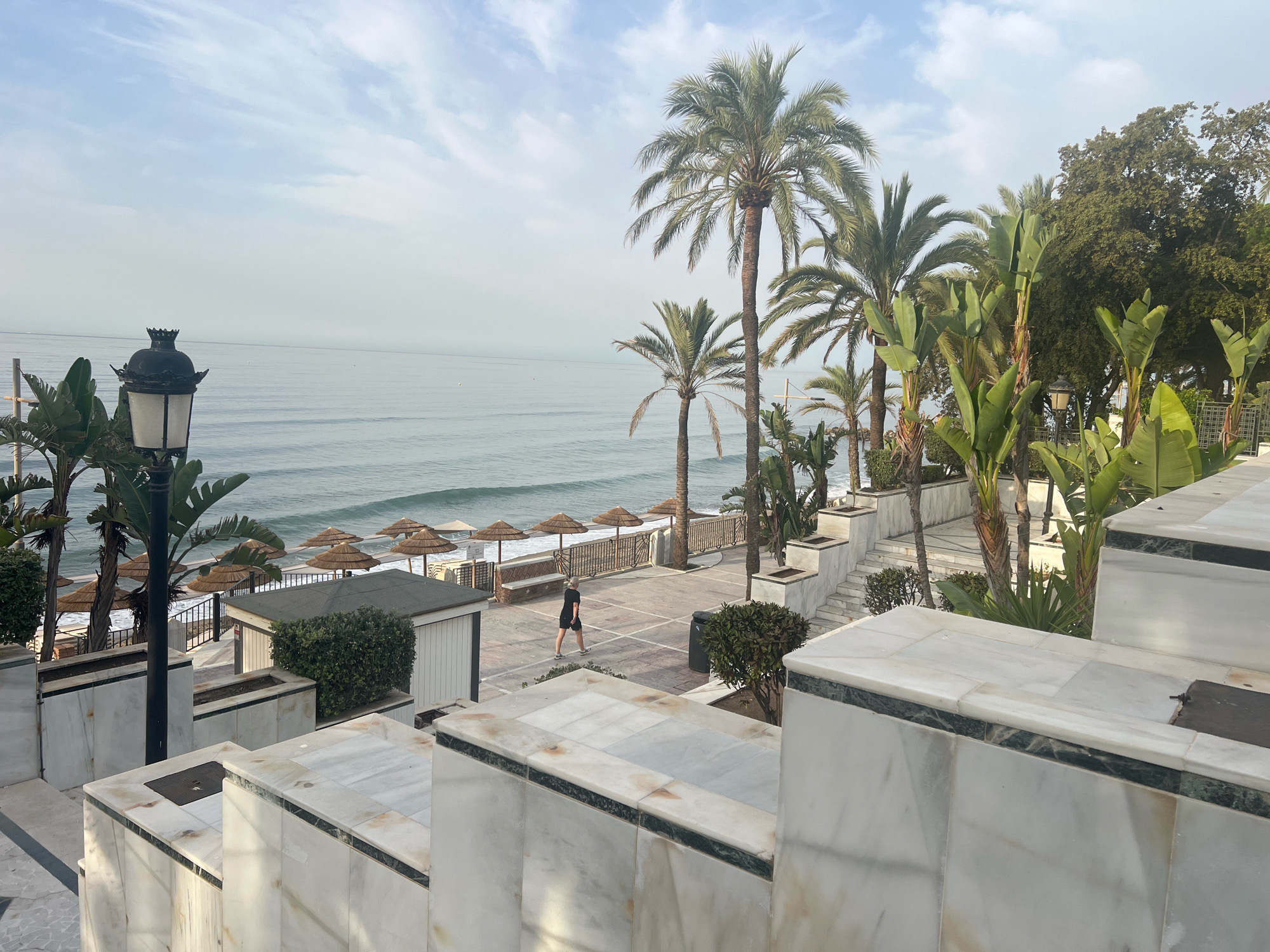 The image depicts a view of the Mediterranean Sea from a promenade in Marbella, Spain. In the foreground, ornate marble steps and planters lead down to a paved walkway. A single person, dressed in dark clothing, walks along the promenade. Beyond the walkway, a row of thatched umbrellas is visible on the beach, with palm trees and lush greenery framing the scene. The sea is calm, with gentle waves rolling towards the shore. The sky is overcast with light clouds. A black, ornate lamppost stands on the left side of the promenade. The scene suggests a tranquil, coastal setting, likely a resort area.