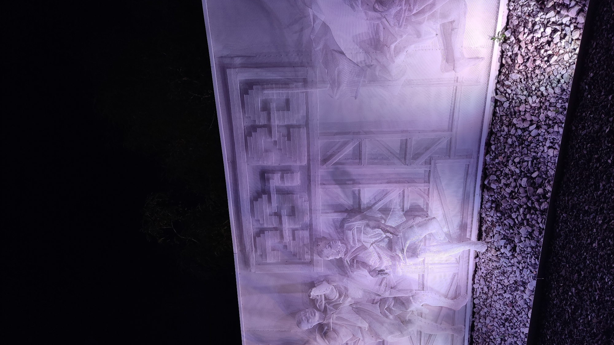 An outdoor sculpture, illuminated with purple light, is visible at night. The sculpture is made of white, mesh-like material and depicts figures that appear to be people in historical clothing. Above the figures, a pattern resembling a maze or a stylized number sequence is visible. The sculpture is situated in front of a dark, unlit background, with some trees visible at the top. The base of the sculpture is lined with small, grey gravel. This artwork is located at the Krasnodar Memorial Museum of Military Vehicles, also known as the Victory Park Museum, in Krasnodar, Russia.