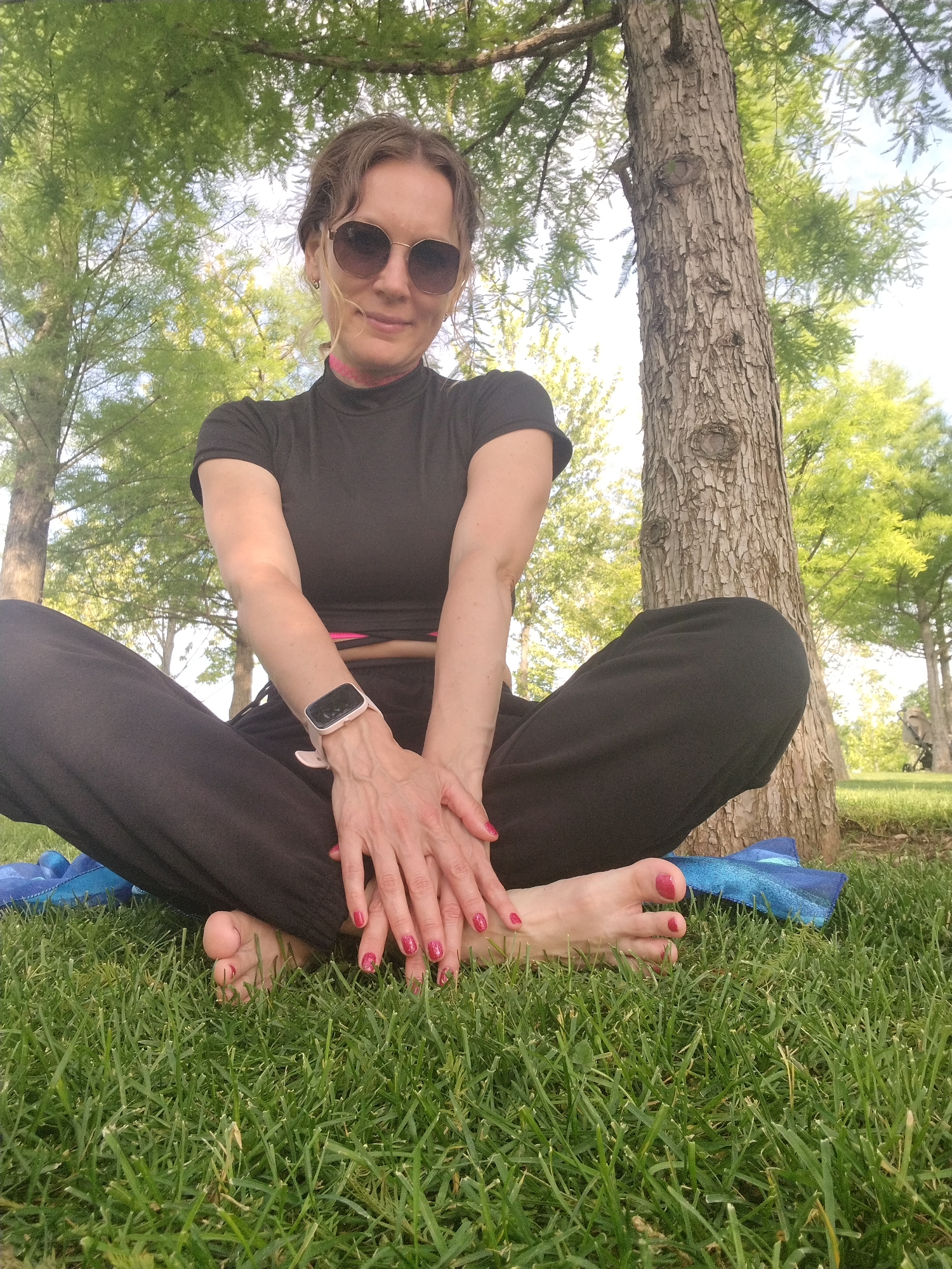A woman sits cross-legged on a patch of grass in a park, leaning against a tree trunk. She wears a black t-shirt and black pants, with a pink choker around her neck and sunglasses. A smartwatch with a light pink band is visible on her left wrist. Her hands are clasped together in her lap, her fingernails painted a glittery pink. Her feet are bare, also showing glittery pink toenails. A blue mat is partially visible beneath her legs. The background features lush green trees and dappled sunlight, suggesting a pleasant outdoor setting. There are no visible landmarks that would specifically place this scene within the city of Krasnodar, Russia.