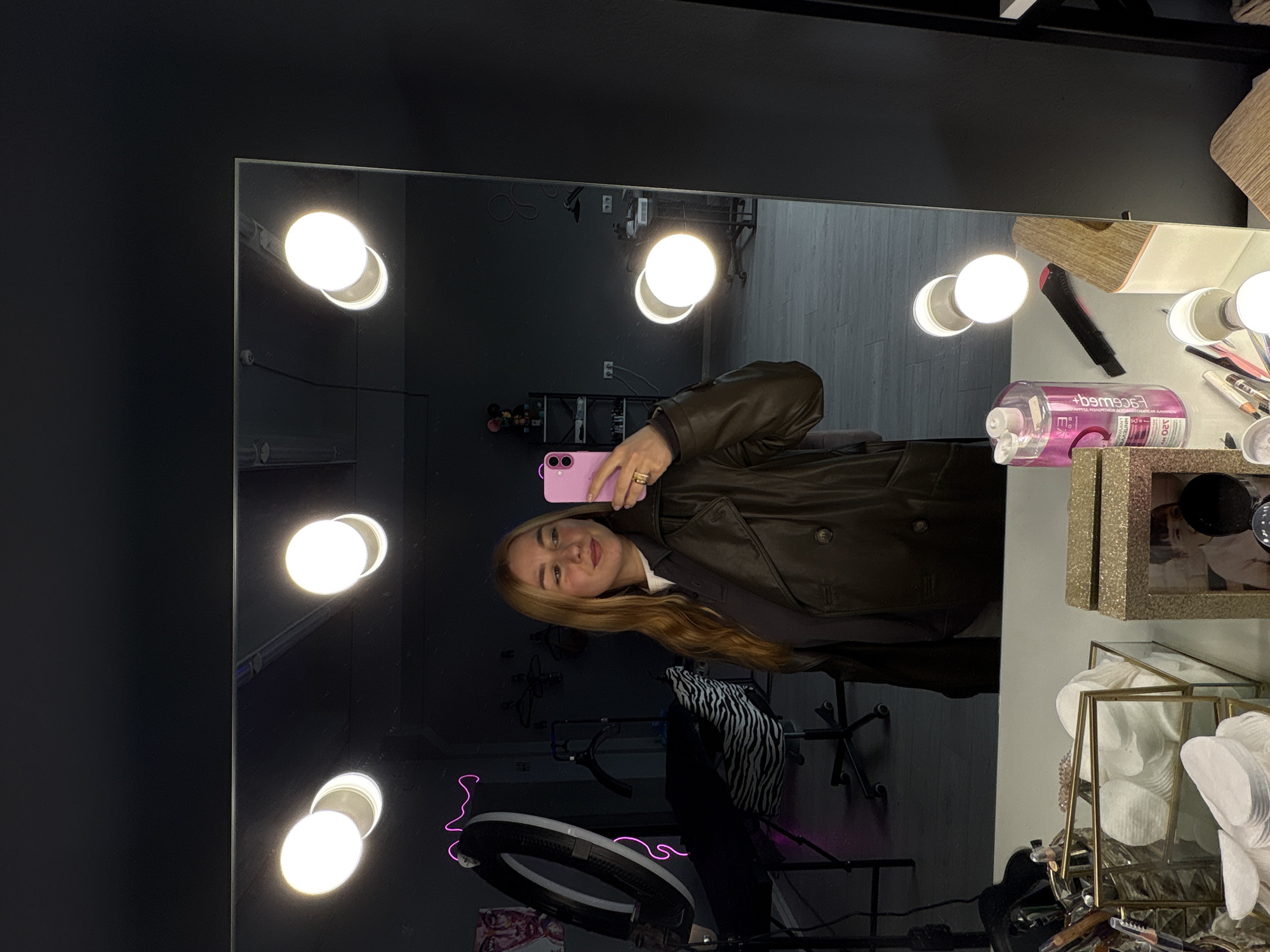 A selfie is taken in front of a makeup mirror with several bulbs. A woman holding a pink cell phone is standing in front of the mirror. The mirror is surrounded by lights. A ring light is visible. The background shows makeup items and a glimpse of the interior of what appears to be a beauty or makeup studio. The location is in the city of Minsk, Belarus.