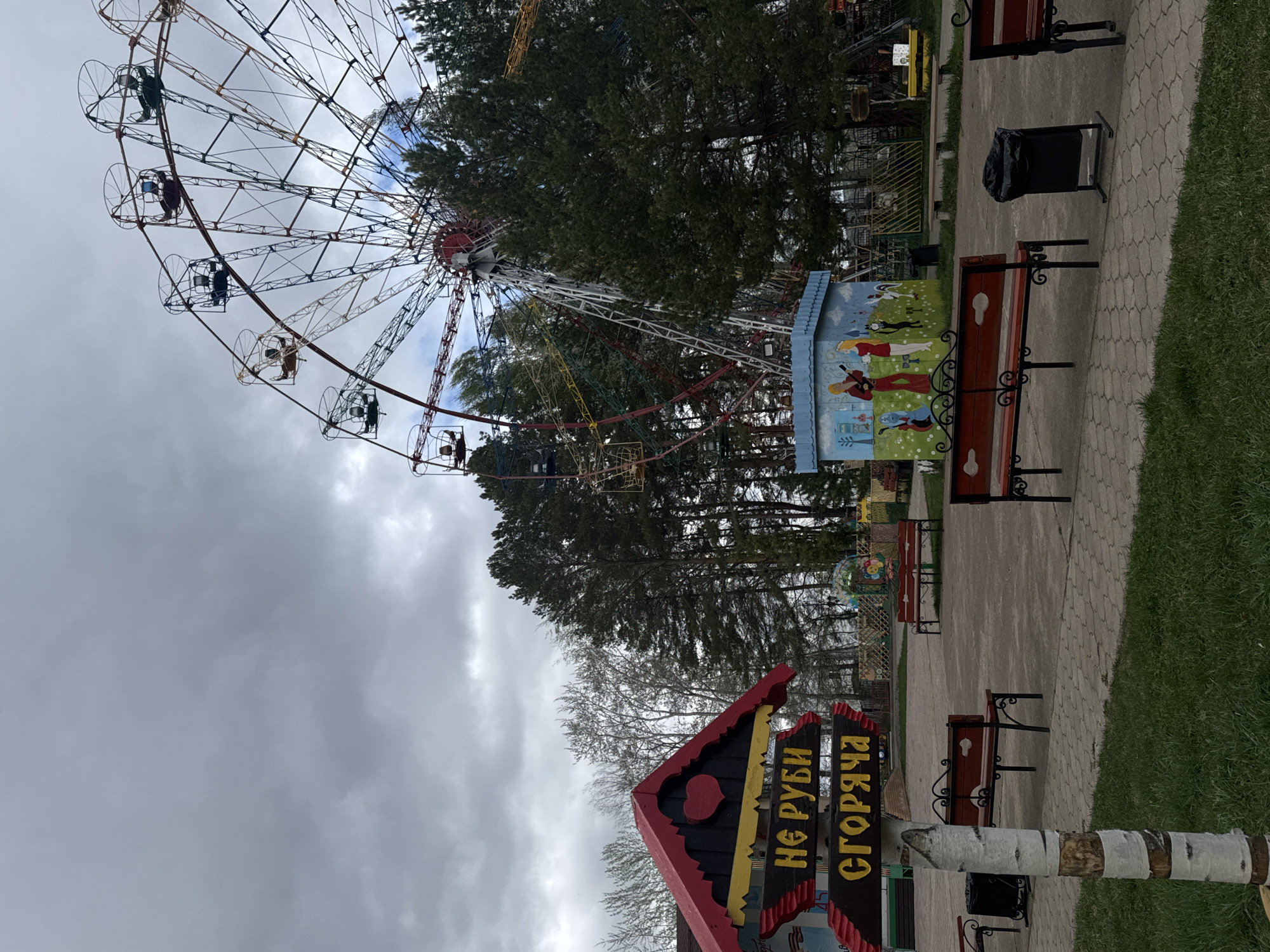 The image depicts an outdoor scene in Chaikovsky, Russia. A Ferris wheel dominates the left side of the frame, with passenger cars visible. Trees form a backdrop behind the Ferris wheel. A paved path runs along the right side of the image, lined with benches and a trash receptacle. In the foreground is a sign with Cyrillic script, mounted on birch logs. The sky is overcast, with a heavy cloud cover. The overall environment is an amusement park or recreation area. No people are directly visible in the photograph.
