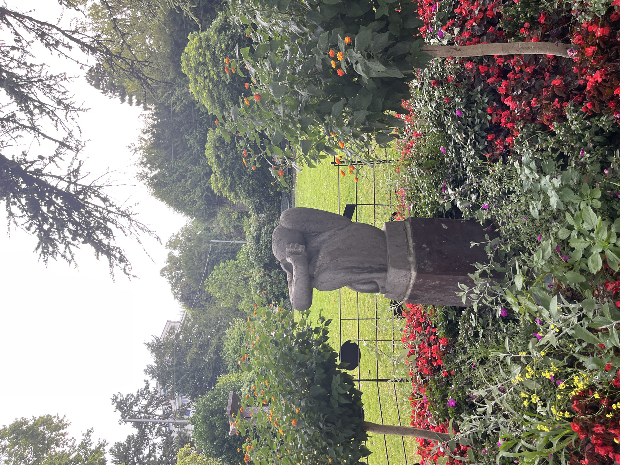 This image depicts a garden scene with a sculpture in the foreground, set against a backdrop of lush greenery and trees. The sculpture, appearing to be made of stone, is of a seated figure with its arms covering its face, resting on a dark rectangular base. In front of the sculpture, a vibrant flowerbed is filled with red and pink blossoms, interspersed with smaller yellow and purple flowers and silvery-green foliage. Two trees with orange flowers are positioned to the left and right of the sculpture, framing the central subject. Beyond the flowerbeds and sculpture, a neatly manicured lawn is visible, bordered by a black metal fence. In the background, dense foliage and trees create a natural setting, with hints of buildings and more distant trees suggesting an urban or suburban environment. The sky is overcast and bright, indicating diffused lighting conditions. This scene is located in the Queen Studio Montreux, Montreux, Switzerland, which is famous for its music history and scenic beauty.