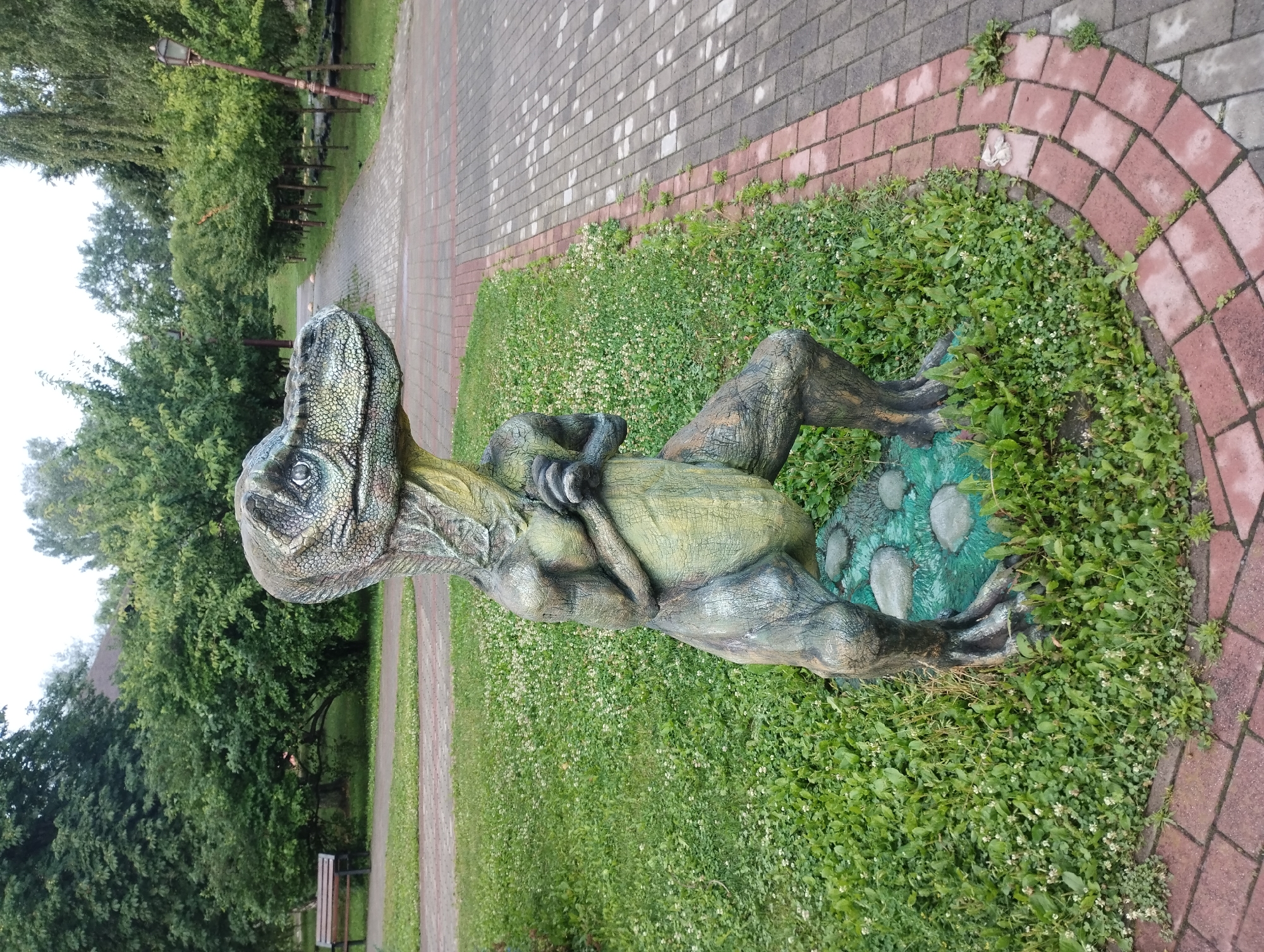 The image shows a statue of a dinosaur in a park. The dinosaur statue is green and brown and is standing on a rocky base surrounded by grass and small white flowers. The park has a paved walkway with red brick edging. Trees and bushes are visible in the background, as well as a park bench and a lamppost. The location is the Sumy City Park in Sumy, Ukraine.