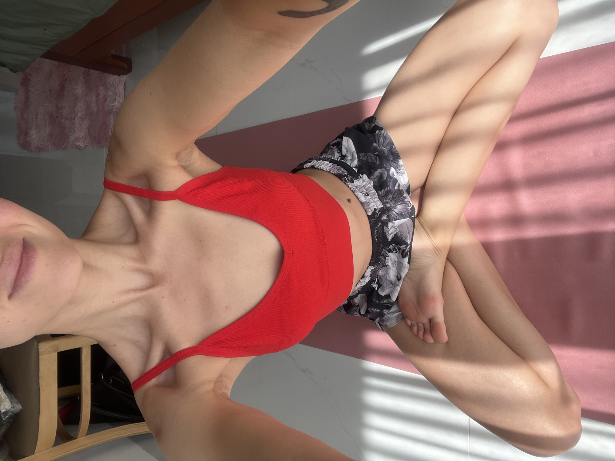 A woman in a red top and black and white floral shorts sits cross-legged on a pink yoga mat on a white tile floor. Sunlight streams through blinds, casting shadows across her body and the mat. The woman's legs are crossed at the ankles, and she is seated on the mat. The scene is indoors. A chair and a fuzzy rug are visible in the background. The image does not contain any references to Nam Natural Heritage Area Trang, Vietnam.