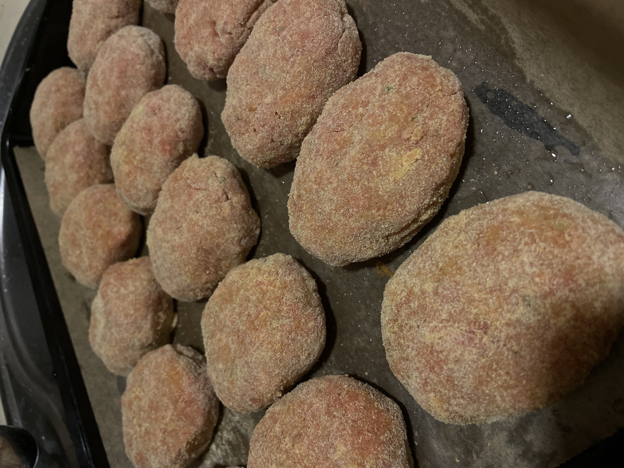 A close-up, overhead shot shows several uncooked, oval-shaped meat patties, each coated in breadcrumbs. They are arranged on a dark baking sheet lined with parchment paper. Some patties are in focus, revealing their pinkish interior and granular coating, while others towards the edges of the frame are softer in focus. The lighting suggests an indoor setting, possibly a kitchen, with a hint of shadow cast by the patties on the parchment paper. One shadow appears to be in the shape of a person. The scene is static, with no visible activity other than the preparation of food. There is no visible text in the image. The location context is La Línea de la Concepción, Spain, which provides a geographical setting for the domestic activity depicted.