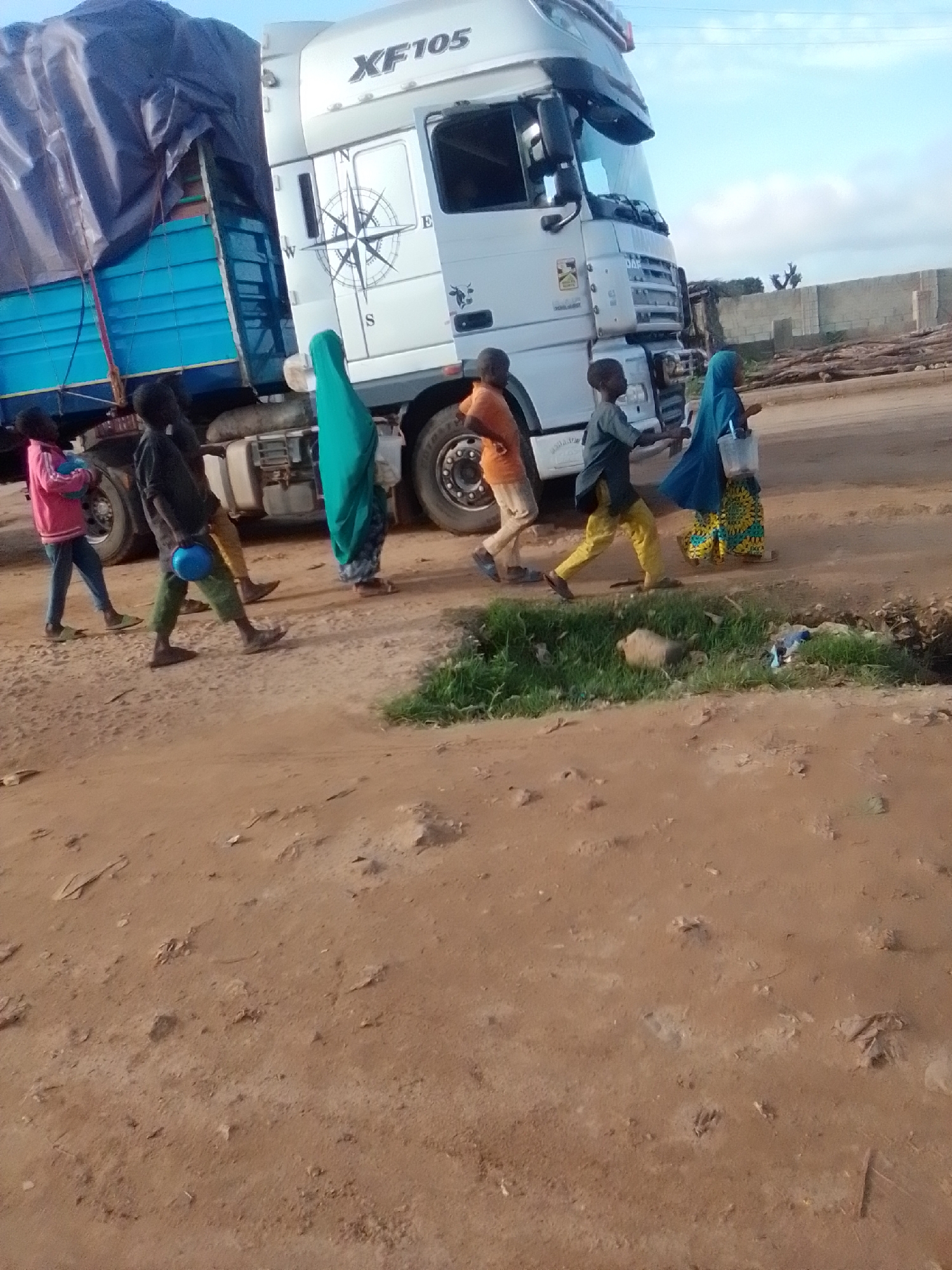 In Kwadon, Nigeria, a white DAF XF105 truck with a blue tarp-covered trailer is parked on a dirt road. Several children are walking near the truck. A girl in a pink jacket carries a blue object. Another child, wearing a green headscarf, walks behind the truck. A boy in an orange shirt and khaki pants is also present, as is a boy in a grey shirt and yellow pants. A girl in a blue headscarf and a patterned dress carries a bucket. The ground is sandy and dry with some patches of grass and debris. In the background, there is a partially constructed wall and some logs. The sky is partly cloudy.