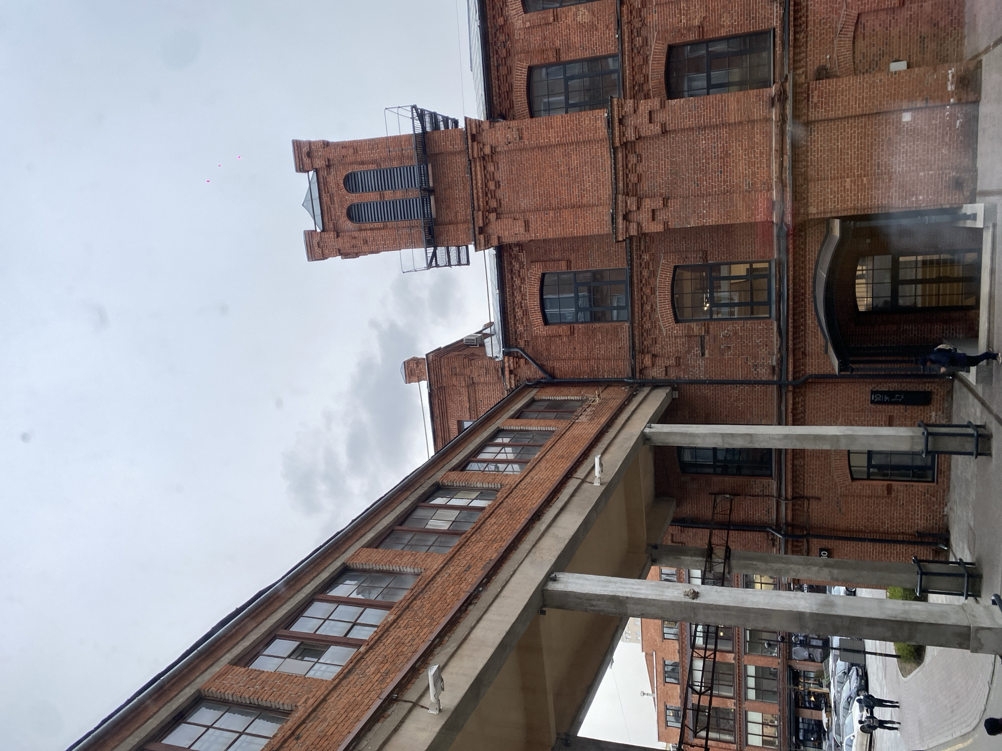 The image displays a complex of red brick buildings connected by an elevated concrete walkway, under a bright, overcast sky in Moscow, Russia. The architecture suggests a repurposed industrial or commercial area.
The main building on the right is constructed of textured red brick with multiple stories and various windows, some arched and some rectangular, fitted with dark frames. A prominent brick tower rises from this section, featuring arched, louvered openings and a metal staircase or railing structure.
To the left, an elongated brick building with an inclined roofline is visible, also featuring numerous windows. This building is linked to the main structure by a substantial concrete walkway supported by robust concrete pillars.
On the ground level beneath the walkway and near the main building, a paved area with several parked cars is visible. At least two individuals are seen walking near an arched entrance of the main building, and another figure is visible further away near the cars.
A notable detail in the sky above the tower are three small, light pink, heart-shaped balloons. Visible text on black signage mounted on the brick wall includes what appears to be "HOK" or "HOW" and a vertical number "2" or "24" near the entrance. Another sign further along the wall reads "194". Through the concrete supports, a black sign with white universal symbols, likely indicating restrooms, can be seen on another building. The time of day is daytime, with a diffused light indicating cloudy weather.
