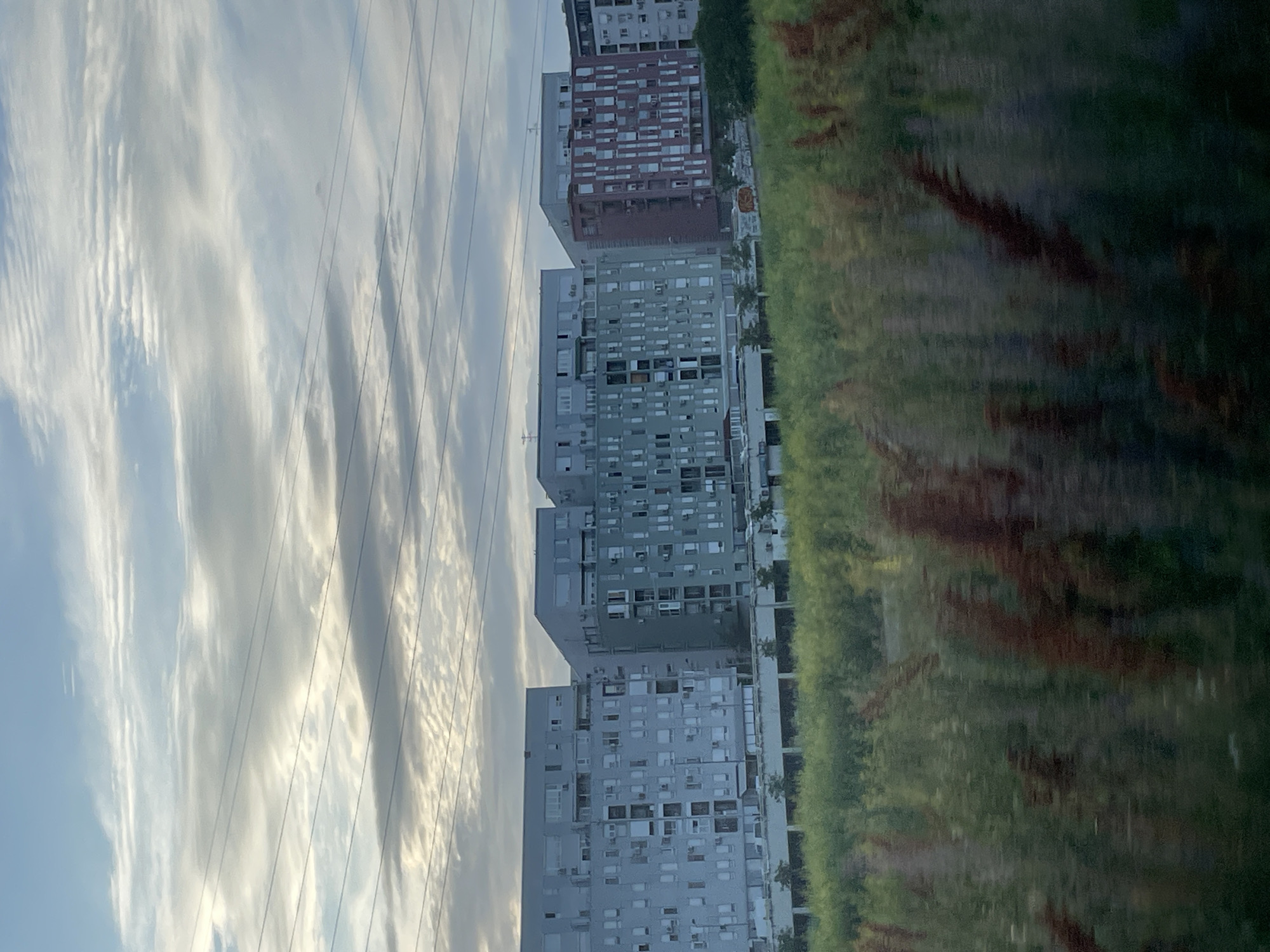 The image shows several tall residential buildings in Belgrade, Serbia, seen against a cloudy sky. Power lines are visible in the upper left. A line of greenery appears on the right side, possibly trees or bushes. The buildings have a modern, repetitive design with many windows. One building has a red brick facade. The overall setting suggests a suburban or residential area within the city.