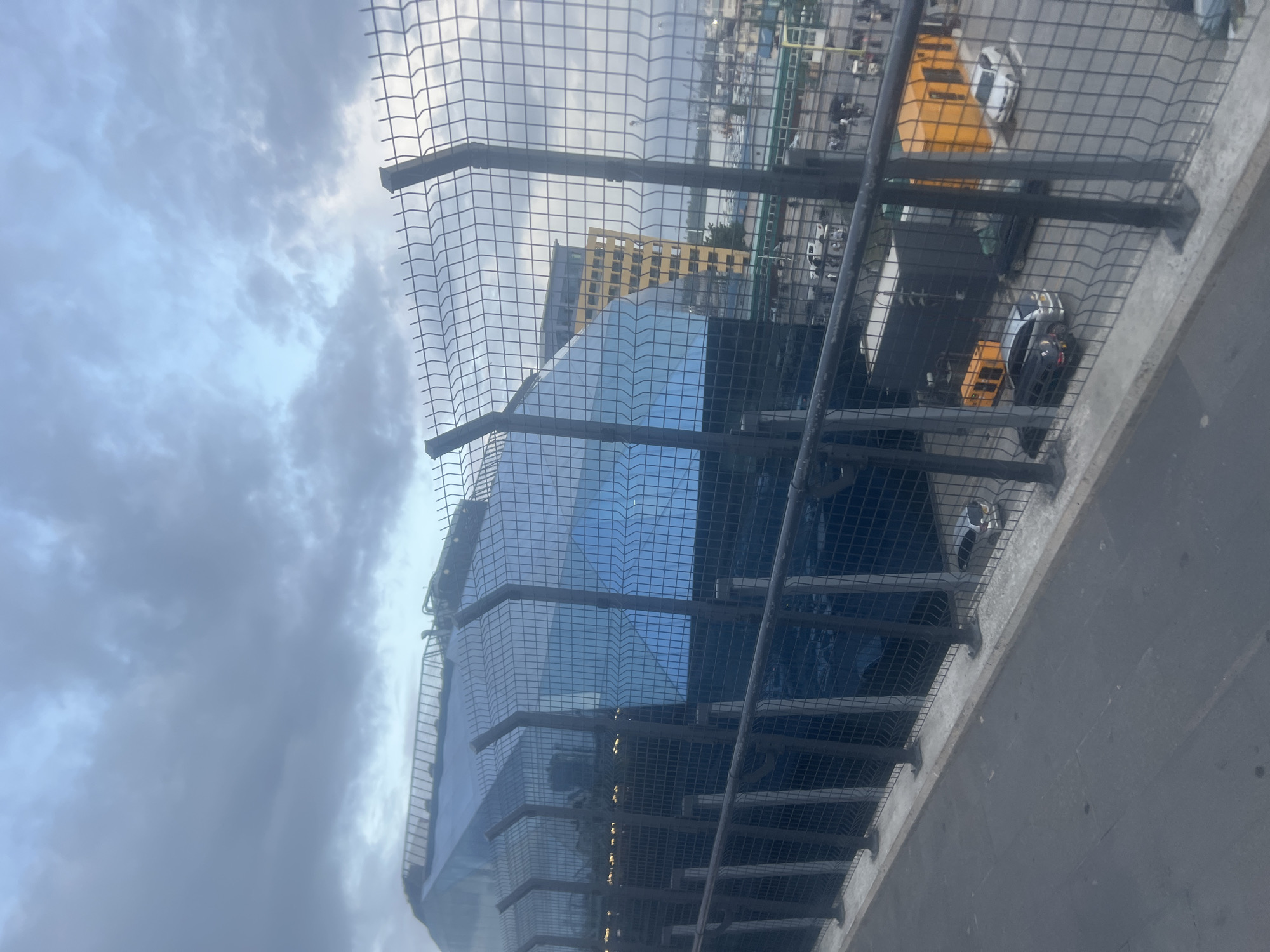 From an elevated perspective through a metal mesh barrier, a modern, high-rise building with reflective glass panels is visible. The building's architecture features a stepped design. In the background, a yellow building is partially visible. Several vehicles, including trucks and cars, are visible, parked or moving on a paved surface in the distance. The sky is overcast with dark clouds. This appears to be the PSPF Twin Towers in Dar es-Salaam, Tanzania.