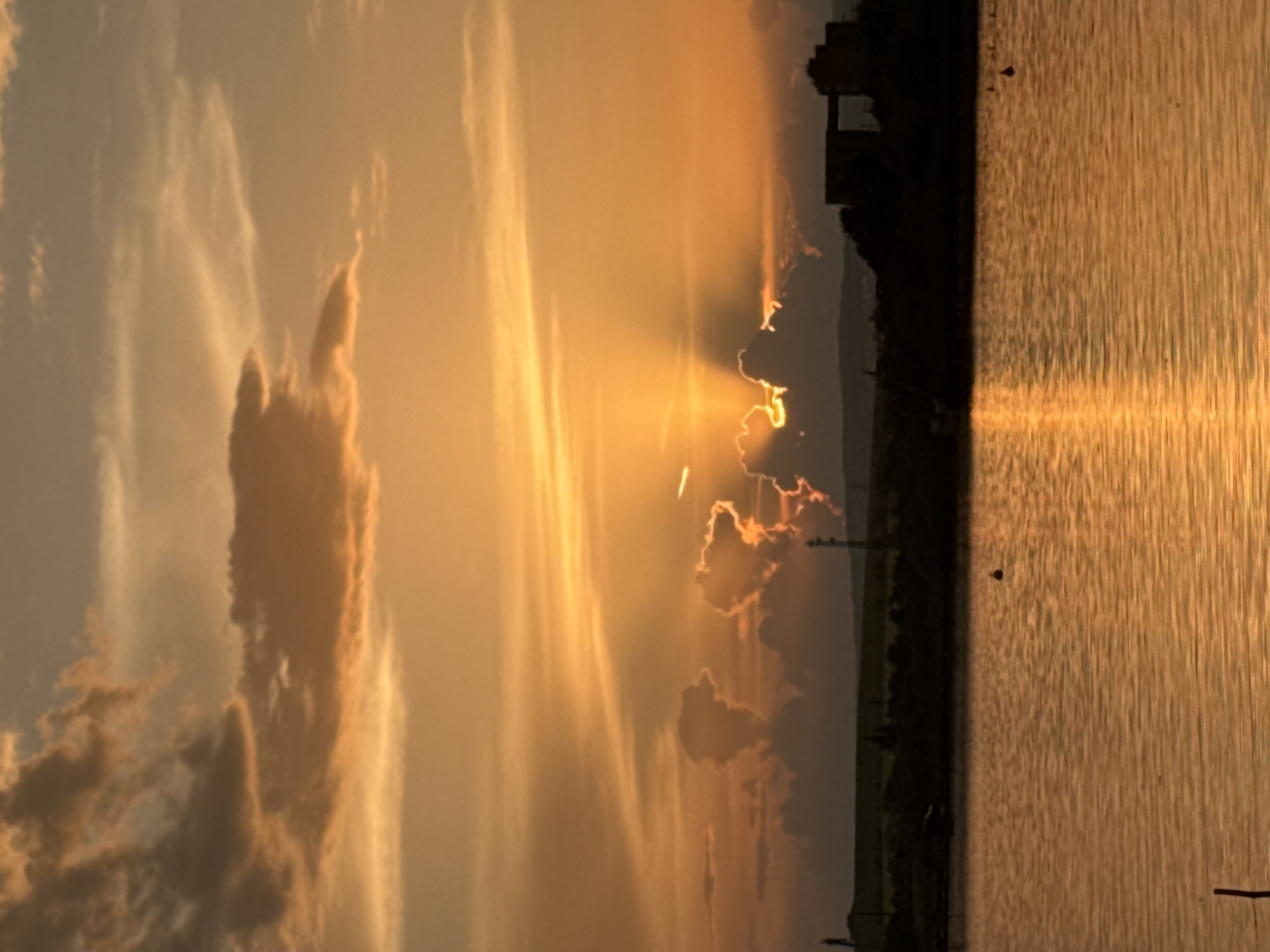 A horizontal, outdoor photograph captures a scene at sunset in Казашко, Bulgaria. The right half of the image is dominated by a body of water, with a reflective surface displaying vertical streaks of orange light. Along the shoreline, a dark silhouette of vegetation and what appears to be a building or structure is visible. The left half of the image showcases an orange sky with prominent clouds. Some clouds are defined, while others are diffuse. Rays of sunlight break through gaps in the clouds, creating a dramatic effect.