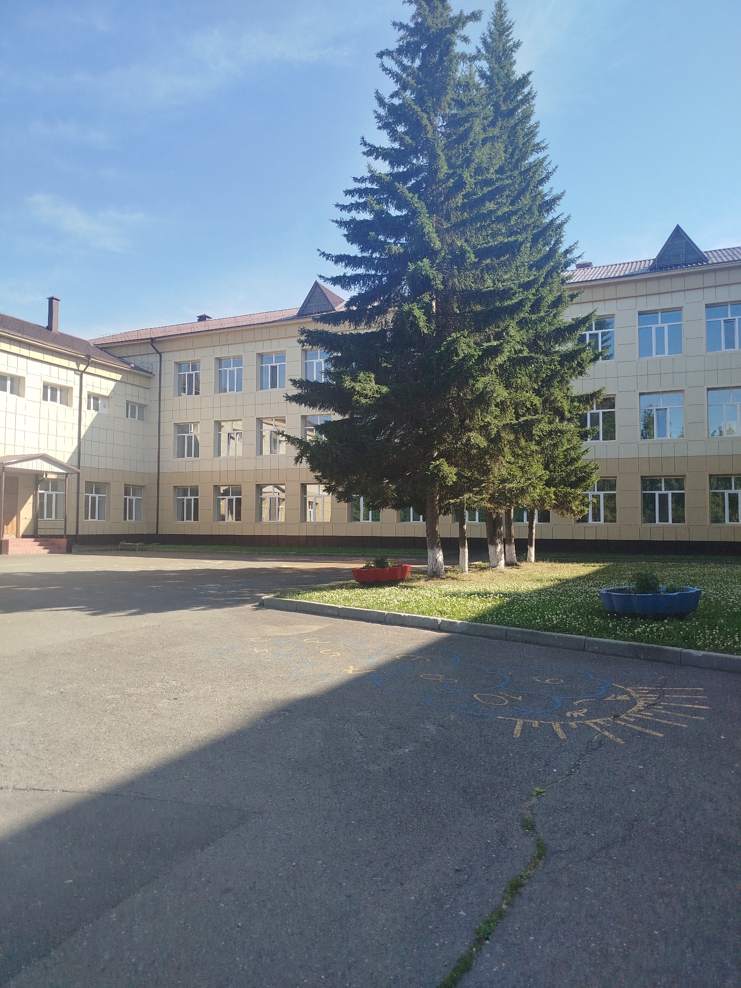 The image depicts the exterior of a two-story school building in Zavodoukovsk, Russia. The building is light beige with a brown roof and numerous windows with white frames. The architecture suggests a functional and modern design. In front of the building, there is a paved courtyard with some painted markings, possibly for games or parking. A strip of green grass borders the courtyard, dotted with small white flowers, and features a few trees, predominantly tall spruce trees. The trees are lush and green, indicating a temperate season, likely spring or summer. Two red planters and a blue trough-like planter are visible, adding some color to the scene. The lighting suggests it is a bright, sunny day with the sun casting a distinct shadow across the courtyard. The sky is clear and blue. There are no people or animals visible in the image. The overall impression is one of a quiet, well-maintained school environment on a pleasant day. The visible text, painted in yellow and blue on the asphalt, appears to be stylized drawings of a sun and possibly letters or numbers, though not clearly decipherable.