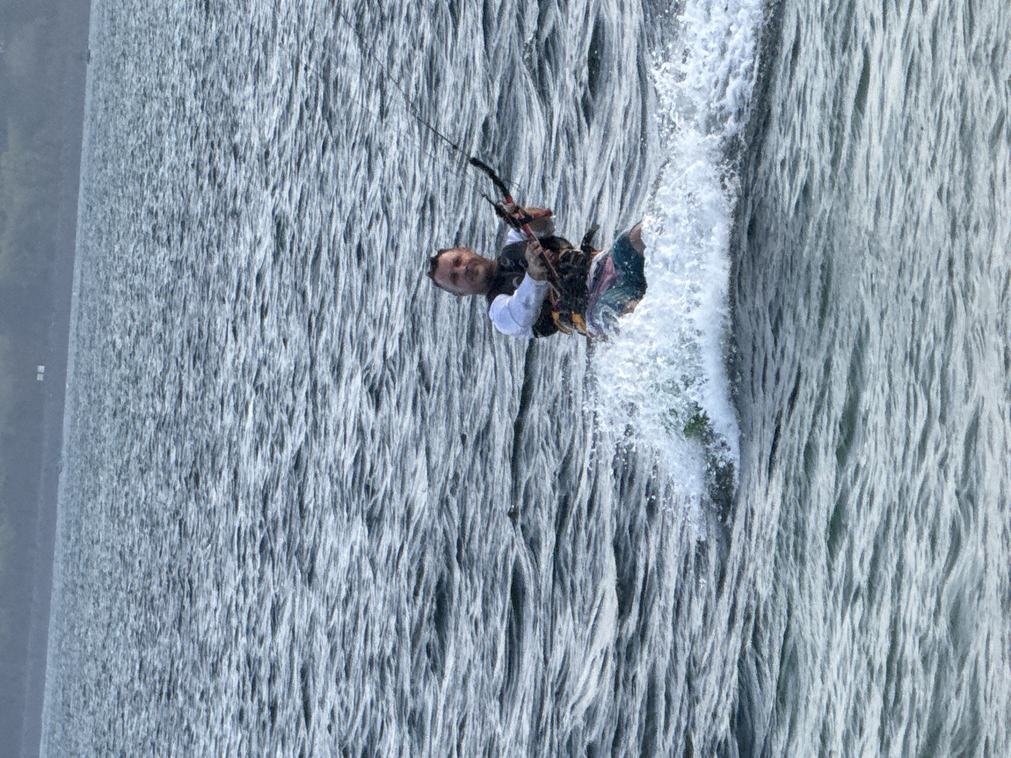 A man is kiteboarding in the ocean. He is wearing a life vest and swim trunks. The water is choppy, and there is a spray of water around him. The background is a blurry image of land. The kite is visible above him, connected by lines. The man is smiling and appears to be enjoying himself.