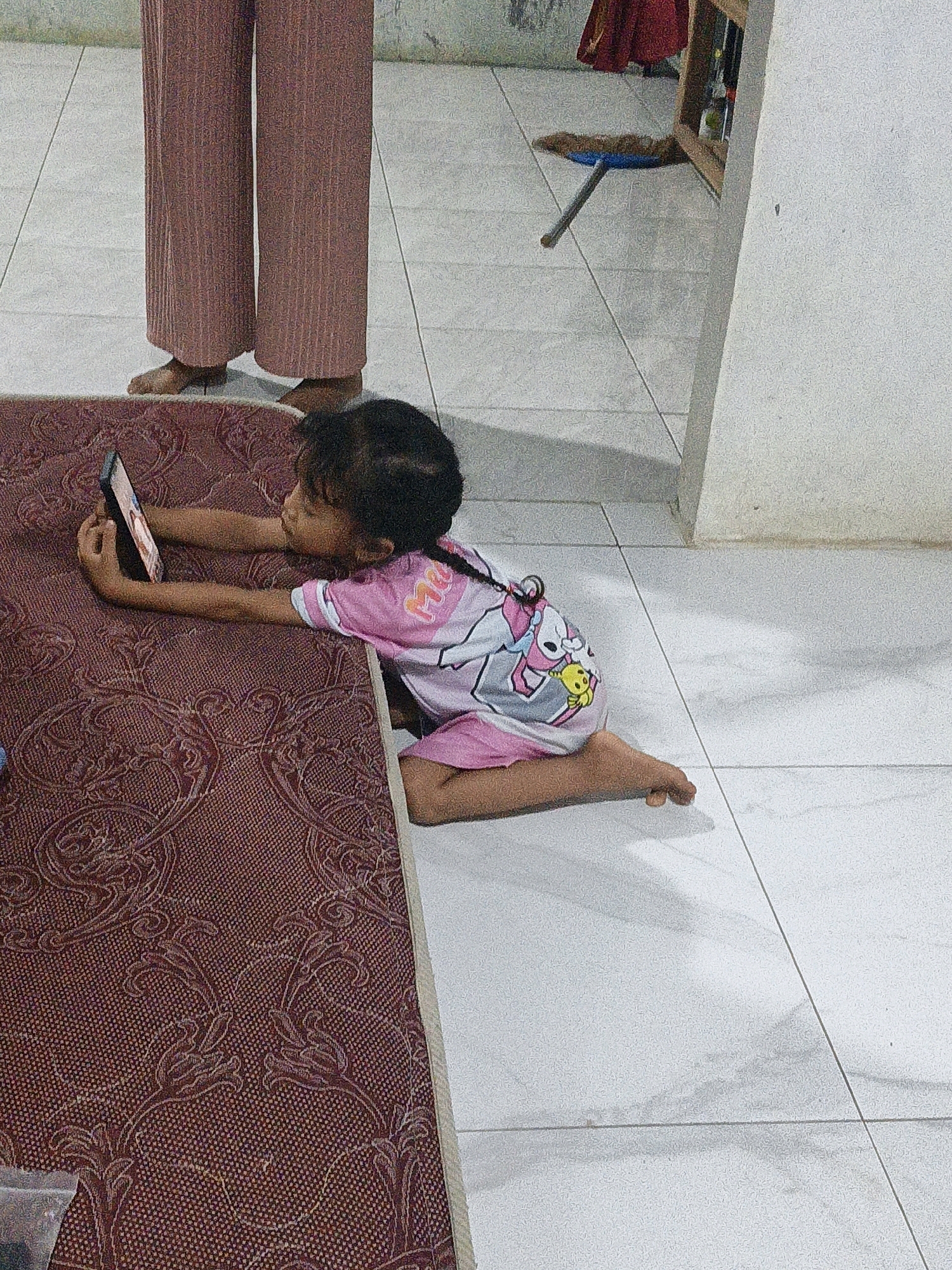 In a domestic indoor setting in Suwawal Timur, Indonesia, a young child lies on their stomach on a maroon patterned rug, engrossed in a smartphone. The child wears a pink t-shirt with white and striped sleeves, and a matching pink skirt. The t-shirt features cartoon characters on the front, including what appears to be a yellow character. The child's hair is dark and pulled back. Their right arm is extended, holding the phone, while their left arm is bent. Their legs are tucked beneath them, with their right foot visible. To the left of the child, the lower legs of an adult are visible, clad in loose-fitting, pleated, rust-colored pants, with bare feet. The floor is tiled in a light color, with distinct grout lines. In the background, a broom with a blue dustpan and a wooden frame are visible, along with a red fabric. The lighting suggests it is daytime, with shadows cast on the floor. The overall scene depicts a moment of quiet personal engagement within a home environment.