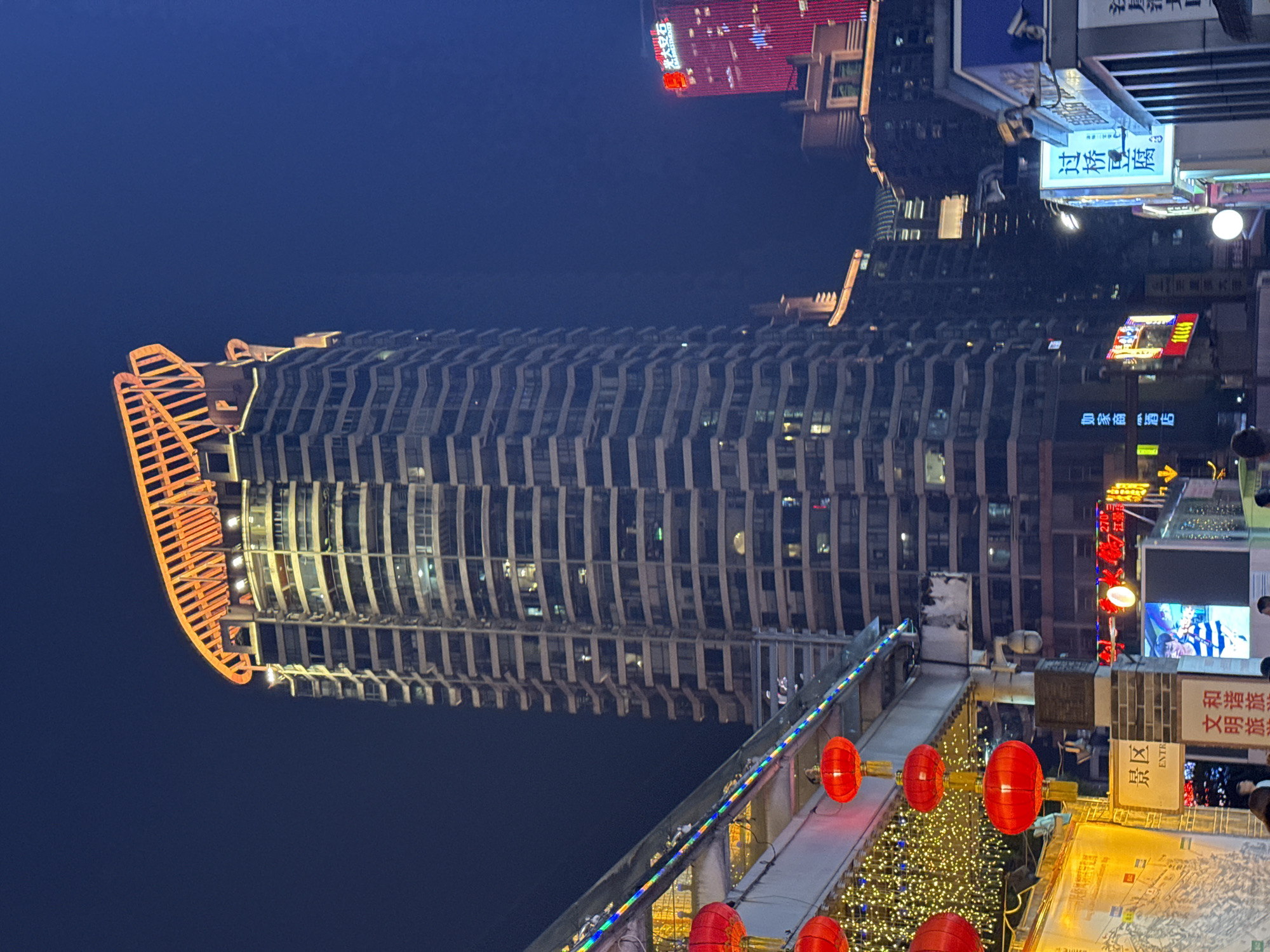 The image depicts a night scene in Yuzhong District, Chongqing, China, featuring the Raffles City Chongqing complex. The most prominent feature is a large, modern skyscraper with a distinctive orange crown-like structure atop. The building's façade has a series of horizontal lines, giving it a layered appearance, and some windows are illuminated.
In the foreground, there is a walkway adorned with red Chinese lanterns, and a Christmas tree with lights is visible. Various signs and buildings with bright lights and neon signs are seen in the background, adding to the urban atmosphere. The image suggests a vibrant commercial district. The sky is dark, indicating nighttime, and overall, the photo showcases a mix of modern architecture and traditional Chinese elements.