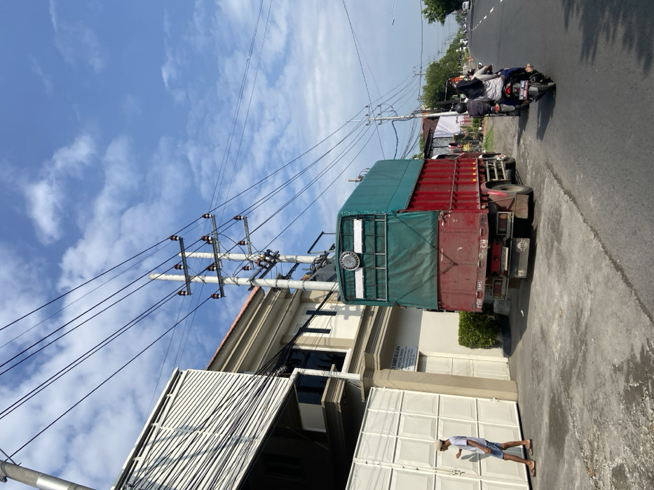 The image shows a street scene in Kecamatan Tanggulangin, Indonesia (latitude: -7.497, longitude: 112.685). A large, red and green truck is parked on the side of the road, facing forward. The truck has a tarp covering its cargo bed and is partially visible through the tarp. A man is standing on the sidewalk near a closed white gate, looking to the left. He is wearing a light blue shirt and shorts. On the other side of the truck, two men are riding motorcycles. The man on the front motorcycle is wearing a black helmet and a white shirt. The man on the back motorcycle is wearing a black helmet and a dark shirt. Both motorcyclists are wearing sandals. The motorcycles are facing forward and are in the middle of the street. On the opposite side of the road, a man is walking away from the camera. Behind the parked truck, there is a two-story building with white walls and a white corrugated metal roof. Power lines and power poles are strung across the street. The day is sunny, with a blue sky and some white clouds. The road surface is paved and there is a sidewalk on the right side of the road. The image appears to be taken from a pedestrian’s perspective.