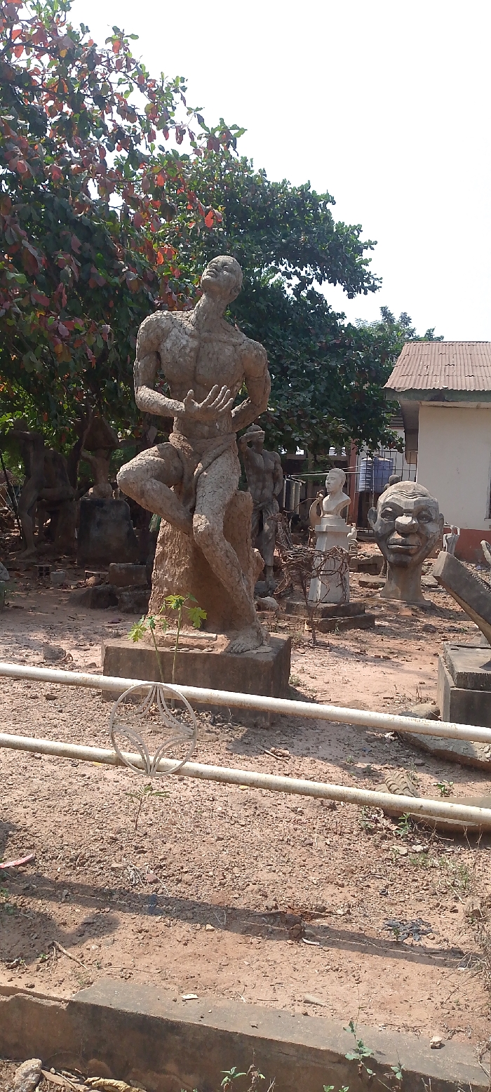 The image depicts an outdoor sculpture garden or workshop area, likely located within the Department of Fine and Applied Arts, University of Nigeria, Nsukka.
In the mid-ground, a prominent, light-colored concrete or cement statue of a muscular male figure is positioned slightly right of center. The figure is depicted in a crouching stance, leaning back on its left leg, with its right knee raised and its left foot resting on a rough, rock-like base. Its head is tilted sharply upward, and its mouth is slightly open, suggesting an expression of anguish, strain, or supplication. Both hands are raised and slightly cupped towards its chest. The sculpture has a coarse, textured surface.
Behind this central statue, numerous other sculptures are visible, varying in size and form. These include other full-body human figures in various poses, some partially obscured by trees, and at least two large human heads, one with exaggerated features positioned to the right. A smaller, white bust of a man is also visible on a pedestal further back. The sculptures appear to be made from similar concrete or cement materials, displaying a uniform weathered appearance.
The ground is predominantly bare, reddish-brown earth with scattered small rocks and sparse green foliage, including a young papaya plant directly behind a low, light-colored metal rail in the foreground. This rail features a circular decorative element with a geometric pattern.
In the background, dense green foliage from large trees, some with reddish leaves, frames the upper left and central portions of the scene. To the far right, a small building with a corrugated metal roof is visible. The sky above is clear and bright white, indicating strong daylight. No human individuals are actively present or performing actions within the frame, only their artistic creations.