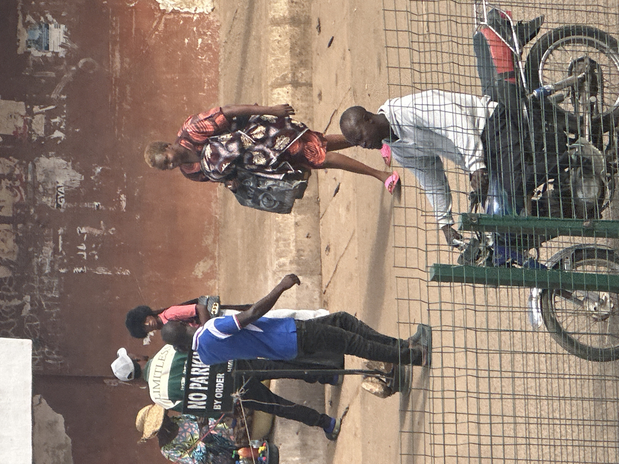 In Bukuru, Nigeria, a scene unfolds on a dusty, outdoor walkway. A young boy with dark, curly hair, wearing a pink shirt, stands near a sign that reads "NO PARKING BY ORDER." To his left, a person in a straw hat and patterned dress is partially visible. Behind them, a man in a white cap and a green and white striped shirt is also present. To the right of the "NO PARKING" sign, a man in a blue shirt and black pants walks away from the camera. Further to the right, a woman with short, blonde hair, dressed in an orange and brown patterned dress, carries a large, dark bag over her shoulder and walks forward. In the foreground, a man wearing a grey hoodie and black pants is seated on a motorcycle, partially obscured by a wire fence. The motorcycle has a black seat with red accents. The environment suggests an urban setting with a weathered, textured wall in the background.