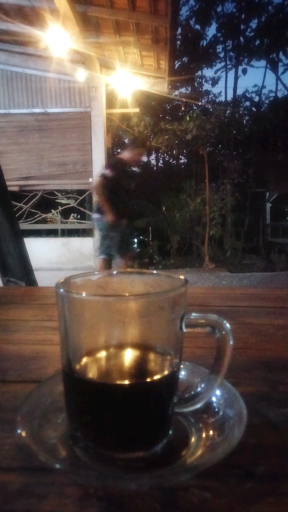 In Plumpang, Indonesia, a glass of dark coffee sits on a wooden table, illuminated by soft overhead lights. The coffee reflects the warm glow of the lights, creating a shimmering surface. In the background, out of focus, a person stands near a building with corrugated metal siding and a wooden roof. Beyond the building, lush green trees and a dark blue twilight sky create a serene outdoor atmosphere. The scene suggests a relaxed evening setting, possibly at an outdoor cafe or a residential area.