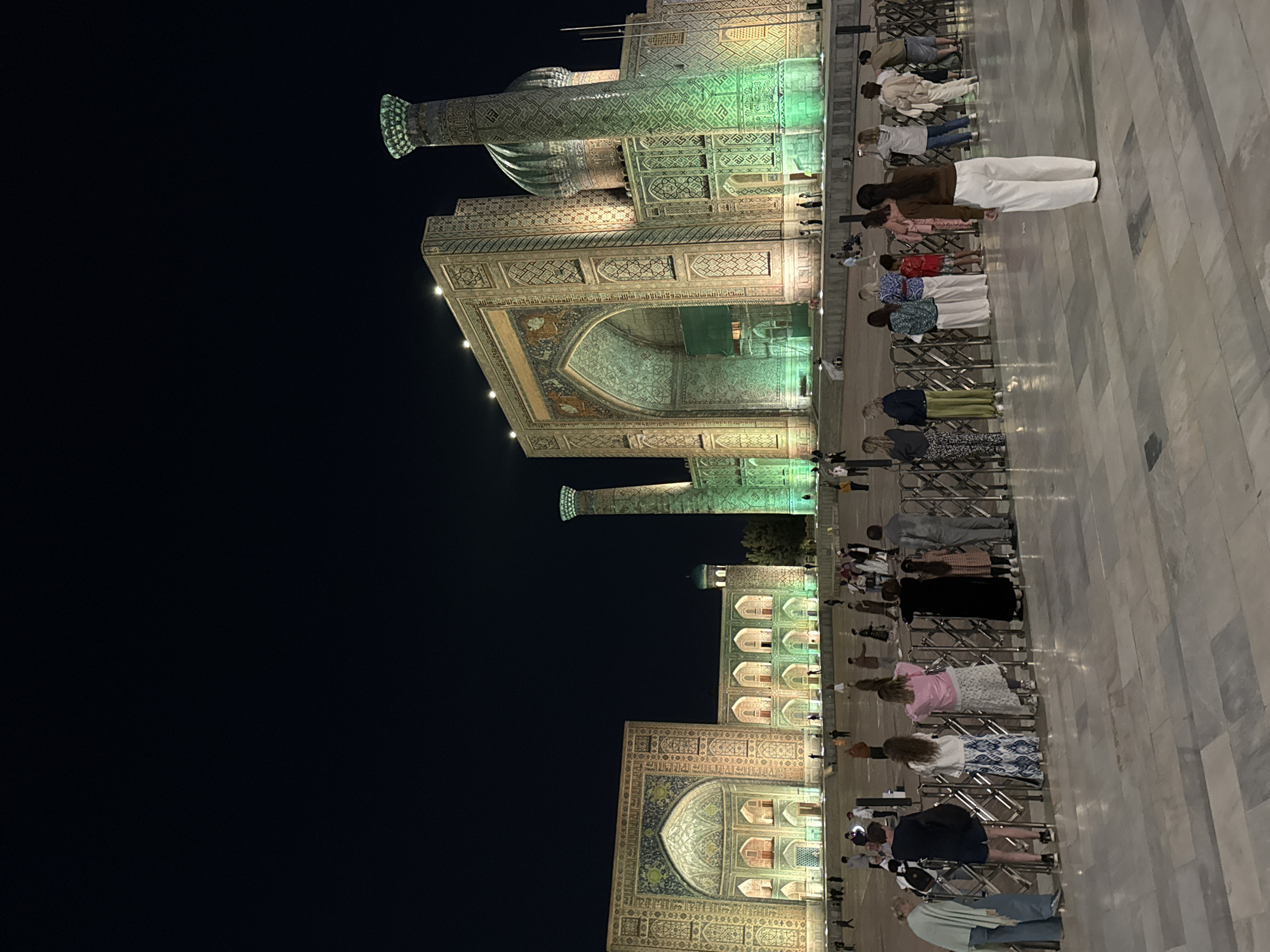 The image shows the Registan Square in Samarkand City, Uzbekistan, at night. Three prominent buildings are visible. People stand in rows, lined up facing the buildings. The buildings and minarets are illuminated with green and white lights.
