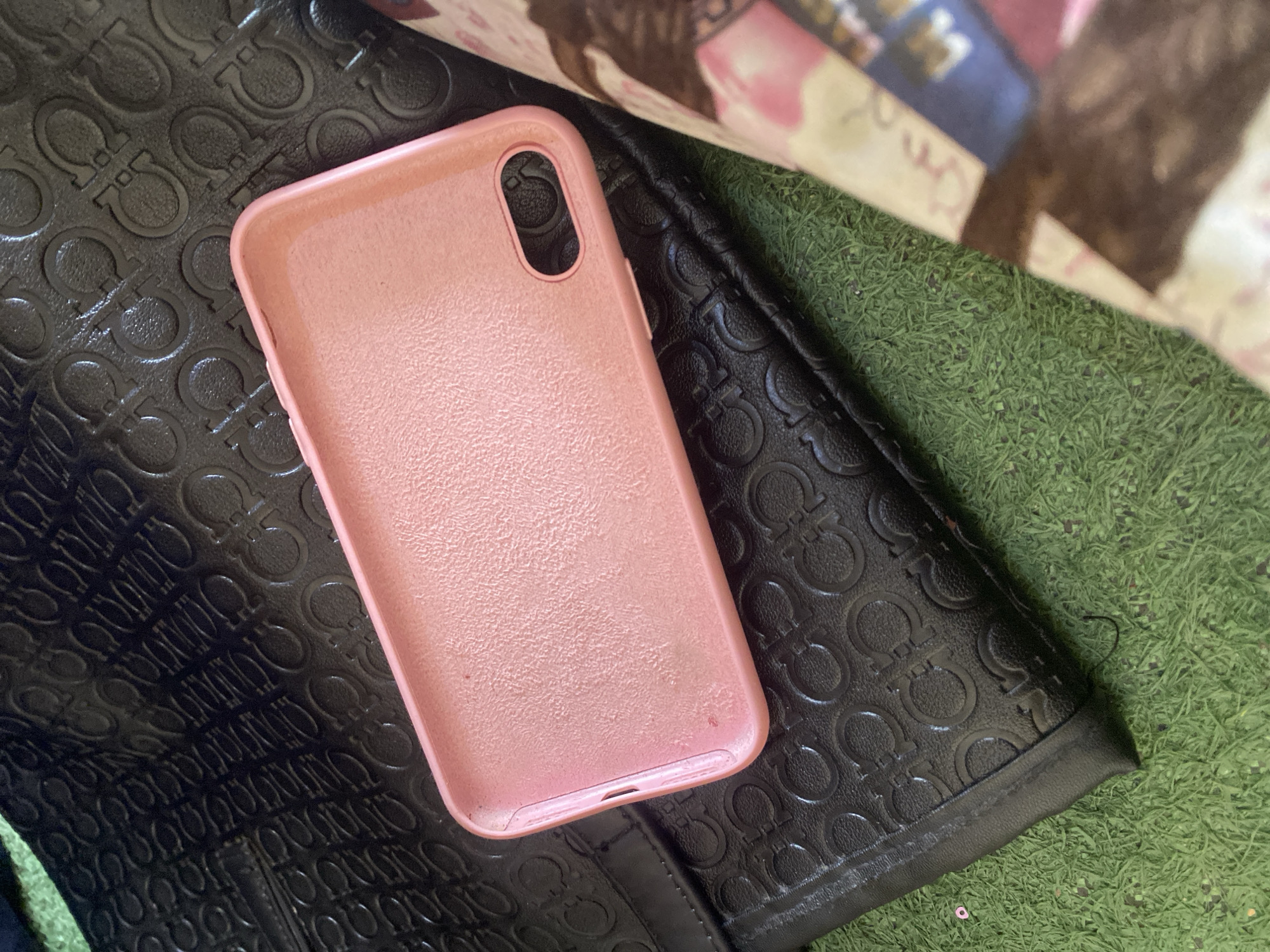 An overhead, close-up shot shows a pink iPhone case laying on top of a black, embossed handbag with an interlocking "G" pattern. The handbag is placed on a green, artificial turf surface. To the right of the handbag, a portion of a magazine or book is visible, featuring a woman's face in the background and colorful patterns. The scene is outdoors, and the lighting suggests it is daytime. The location is specified as Jauro Yinu, Nigeria.
