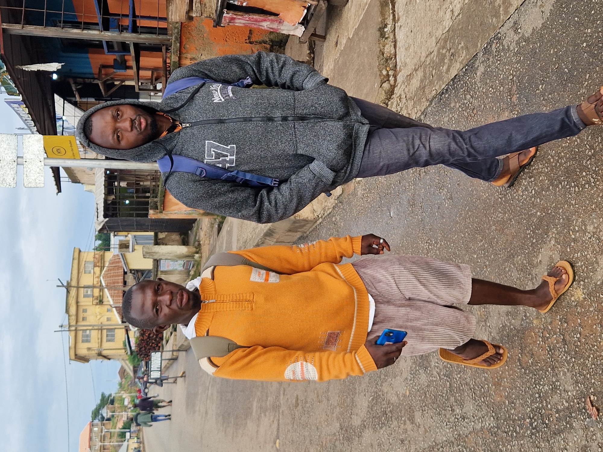 In Kabong, Nigeria, two men walk down a paved street under an overcast sky. The man on the left wears an orange and beige patterned sweater with a white collar, tan corduroy shorts, and brown flip-flops. He carries a beige backpack and holds a blue smartphone in his right hand. The man on the right wears a gray hooded jacket with white lettering, dark blue jeans, and brown sandals. He also has a blue backpack slung over his shoulders. Buildings with orange and blue facades, and some with visible construction materials, line the street. In the background, several other people are visible walking further down the street, and a yellow building stands on a hill.
