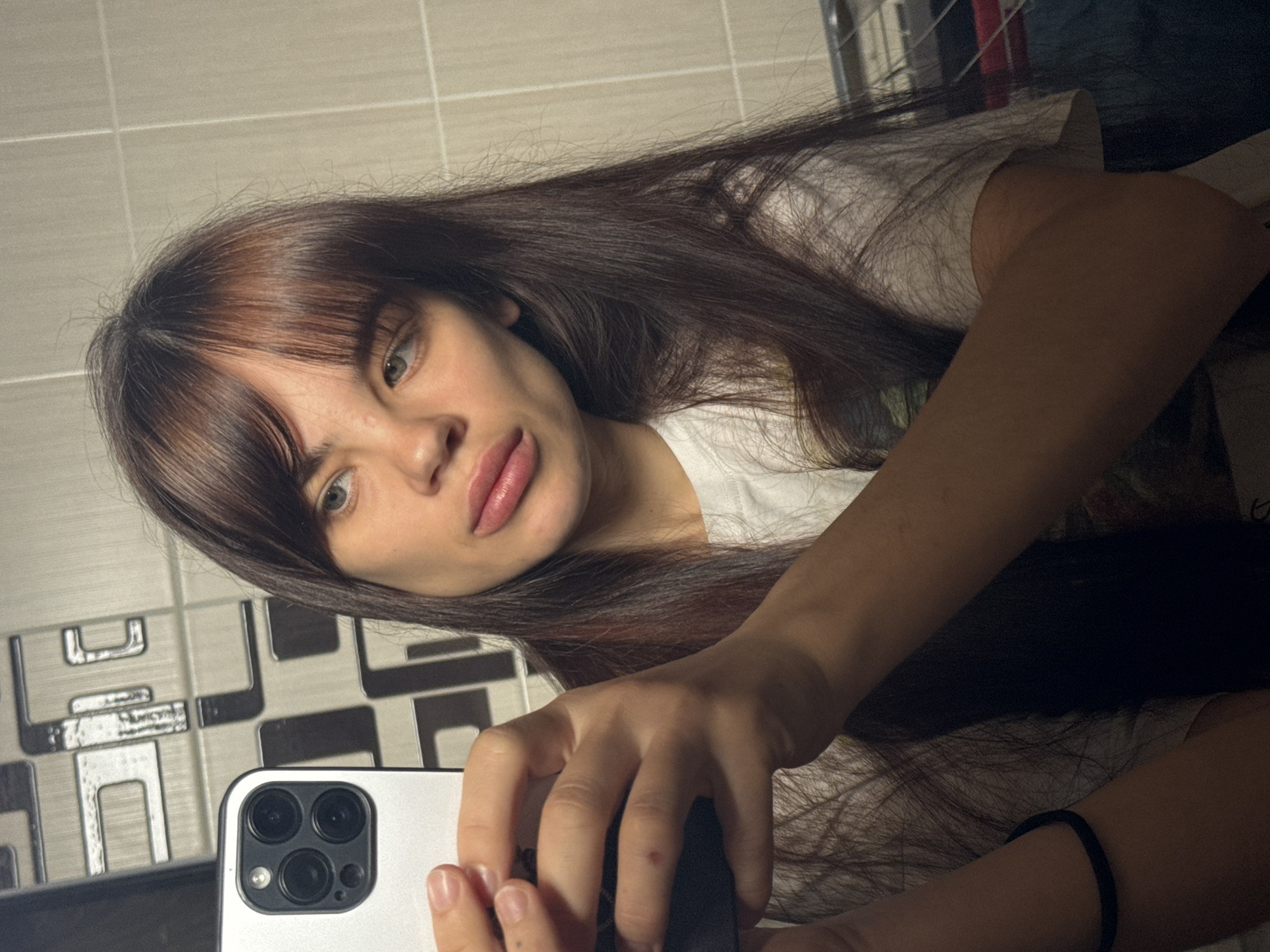 A woman with long brown hair and bangs is taking a selfie. She is holding a silver-colored iPhone in her right hand. She is wearing a white t-shirt and a black elastic bracelet on her left wrist. The background appears to be a bathroom with patterned gray and black tiles. The location is unknown.