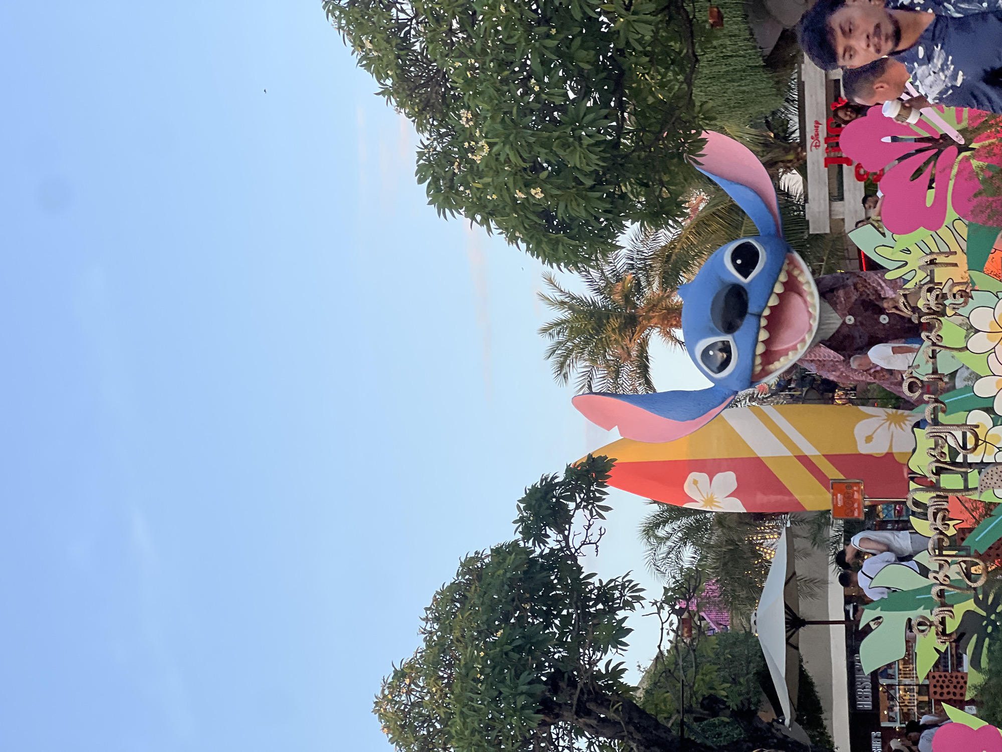 This image captures a daytime scene in Kuta, Indonesia, likely at an outdoor entertainment venue or resort, given the presence of a large Stitch character display. The Stitch figure, a popular Disney character, is a prominent blue, standing with a large, open mouth and wide eyes. It is dressed in a brown patterned jacket. Behind Stitch, a vibrantly colored surfboard featuring hibiscus flowers is propped up. In the background, various trees with lush green foliage and some flowering trees are visible, along with palm trees. The sky is a clear blue with a few wispy clouds.
In the foreground and midground, several people are present. To the left, a white umbrella shades a section of the area, and a store sign that reads "HERSHEY'S" is visible. Two men are seen standing near the Hershey's store, appearing to be conversing. Further to the right, a man in a dark patterned shirt is in the foreground, looking towards the right of the frame. Other individuals are scattered throughout the scene, some appearing to be shoppers or visitors. The overall environment suggests a lively, tropical setting, possibly a shopping or recreational area. The presence of the Disney character and surfboard suggests a family-friendly or tourist-oriented location.