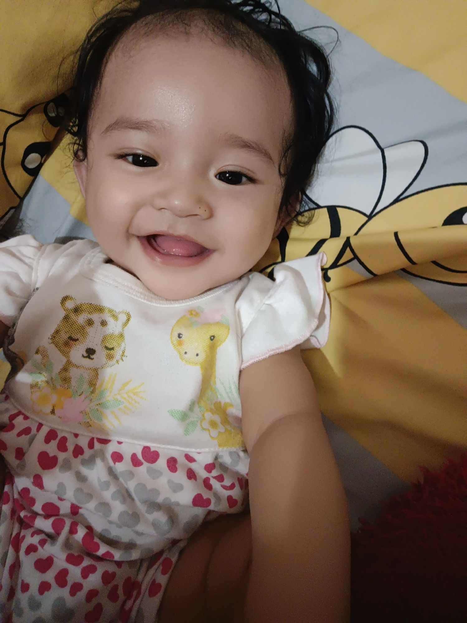 An infant is lying on a yellow and gray patterned blanket. The infant is wearing a white onesie with cartoon animals and hearts on it. The infant is smiling and looking at the camera. The background shows a part of a bed and a pillow with a bee design. The location is indoors in Kawua, Indonesia.