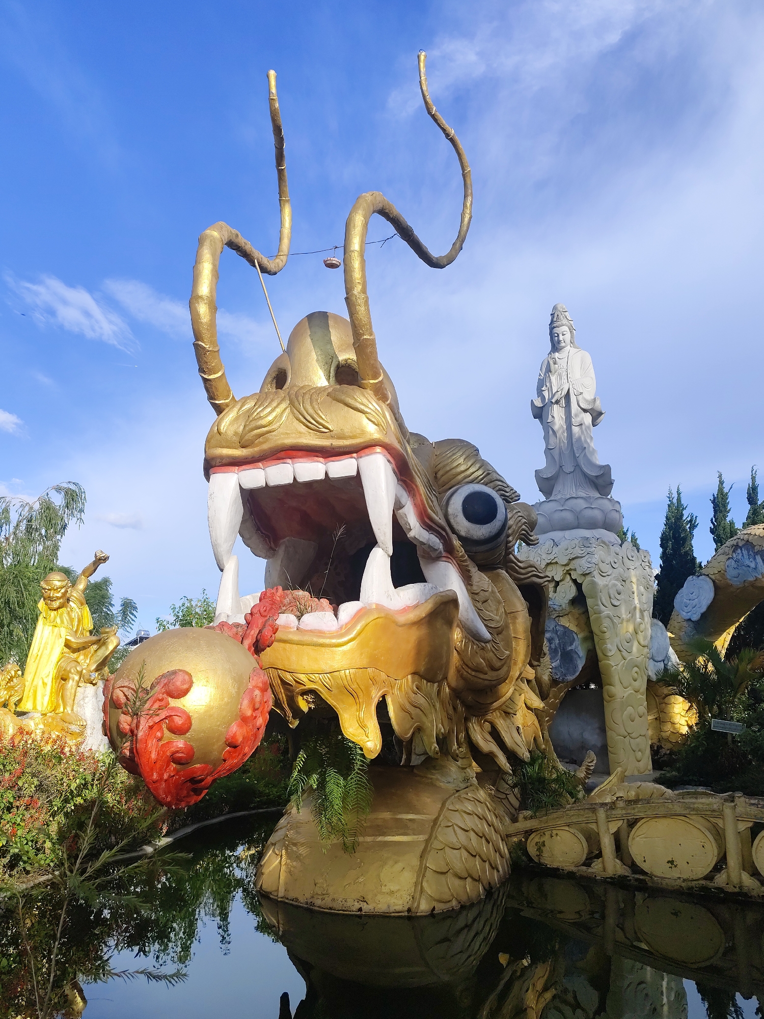 This media file captures a vibrant and ornate outdoor scene, likely at a temple or religious site in Da Lat, Vietnam. The primary subject is a large, golden dragon sculpture, its mouth open in a fearsome display, with sharp white teeth and a red tongue. The dragon is positioned in the foreground, partially submerged in a dark body of water that reflects the sky and surrounding structures.
Behind the dragon, a serene white statue of a standing figure, possibly a deity like Guanyin, is visible atop a sculpted, cloud-like base. To the left of the dragon, another golden sculpture of a seated figure in flowing robes can be seen, adding to the rich visual tapestry of the site. The scene is surrounded by lush greenery, including trees and shrubs with red flowers, enhancing the natural beauty of the setting.
The sky above is a clear blue with wispy white clouds, suggesting it is daytime, likely late afternoon given the warm lighting that casts gentle shadows. The overall atmosphere appears peaceful and awe-inspiring, a common sentiment evoked by such elaborate religious art and natural landscapes. There is no visible text in the image.