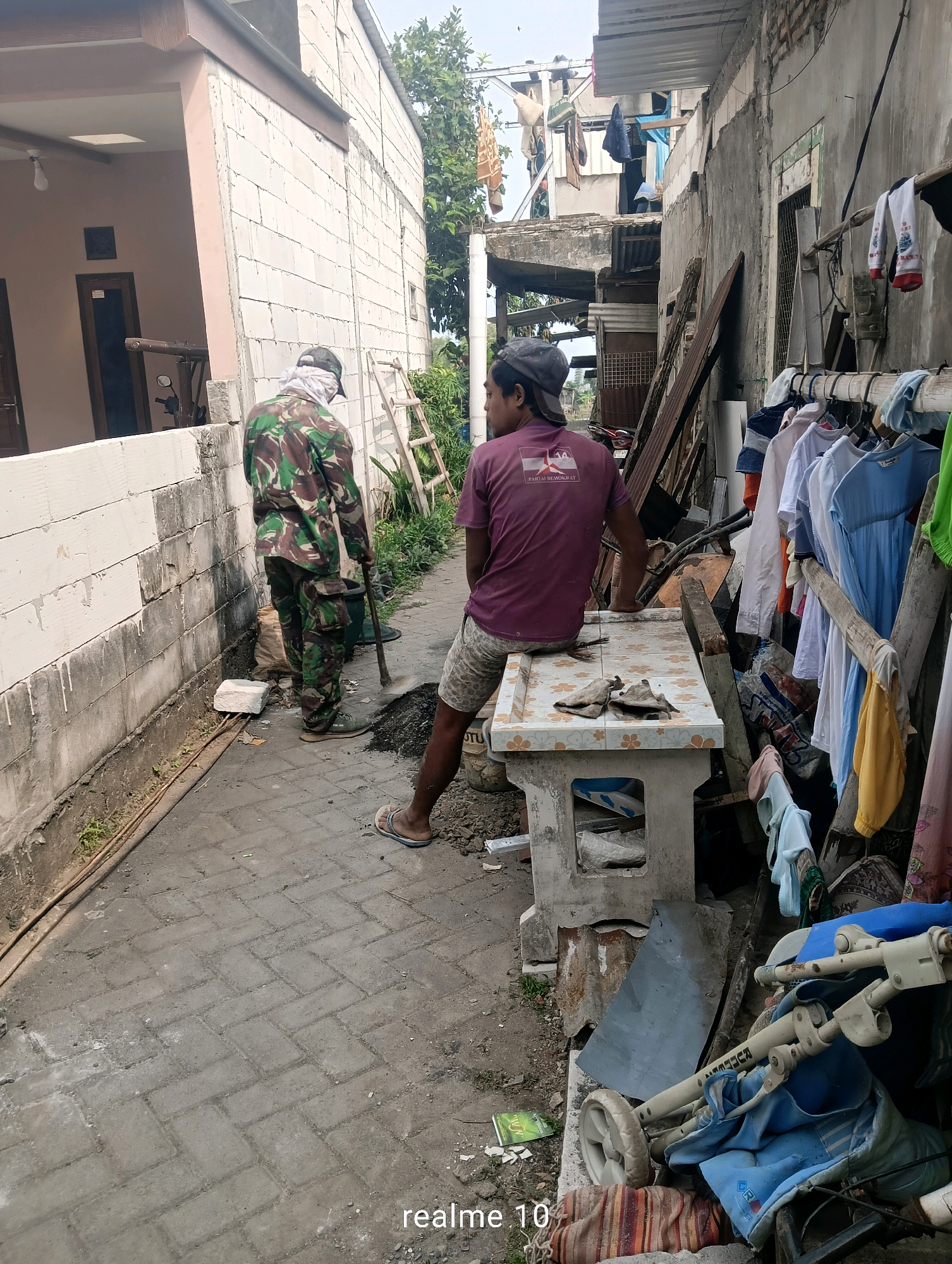 In the city of Batu, Indonesia, a man in camouflage fatigues works with a shovel and bucket on a brick-paved path. He is near a white brick wall and a house with a light-colored facade. To his right, another man sits on a low, tiled table, wearing a purple t-shirt and patterned shorts. A clothesline with various garments hangs on a wooden structure to the right, and a baby stroller is visible in the foreground, partially covered by a blue fabric. The environment appears to be a narrow alley or walkway between buildings, with some debris and overgrown vegetation present.