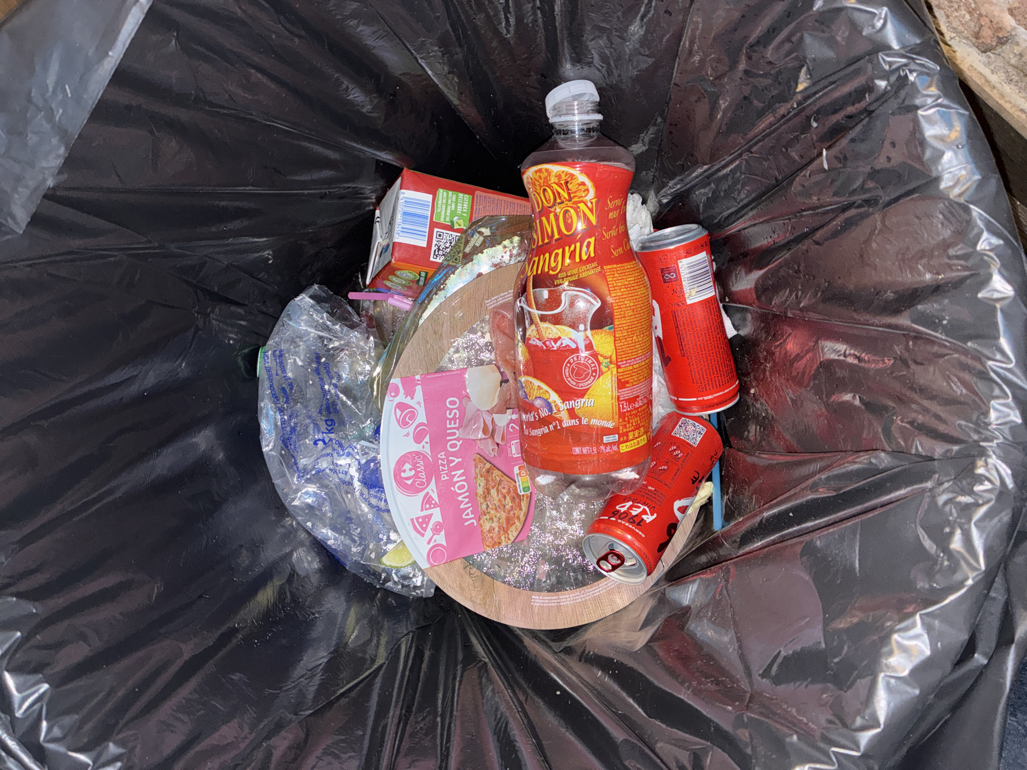 An overhead shot captures the contents of a black trash bag, filled with various discarded items, suggesting a recent meal and beverage consumption. The scene is brightly lit, indicating daytime.
The most prominent item is a 1.5-liter plastic bottle of "DON SIMÓN Sangria," a "RED WINE COCKTAIL," featuring text in Spanish, French ("La Sangria nº1 dans le monde"), English ("World's No. 1 Sangria"), and Japanese. The label also specifies "7% alc/vol." Next to it are two red aluminum cans, one clearly visible as "1906 RED," likely a Spanish beer brand, with "CERVEZAS" visible.
Also present is packaging for a "Classic PIZZA JAMÓN Y QUESO" (Ham and Cheese pizza), with a circular, wooden-textured base underneath it. Another red cardboard carton, partially obscured, shows a barcode and text like "DESCUBRE NUESTROS PRODUCTOS" and "ESCANEA." A clear plastic bag contains some plastic straws and what appears to be discarded lime or lemon peels. The items collectively suggest a gathering or meal in Barcelona, Spain, given the Spanish branding and multicultural labeling on the sangria. All items appear to be empty or used, consistent with their disposal in a trash bag.