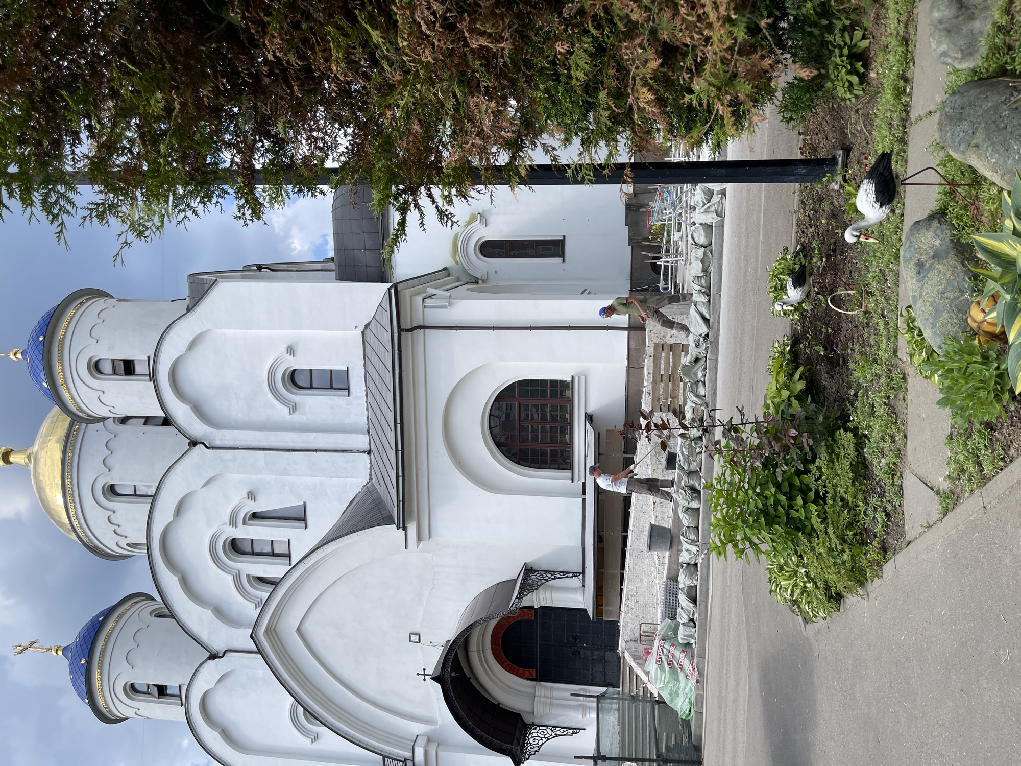 In Moscow, Russia, a white Orthodox church with blue and gold domes stands under a cloudy sky. Two men are working on landscaping and construction in front of the church. One man, wearing a blue cap and green shirt, is raking the ground near a pile of sandbags. The other man, wearing a white shirt and a gray cap, is also raking. In the foreground, a garden bed features lush green plants, decorative rocks, and two stork statues. Construction materials, including sandbags and green plastic sheeting, are visible near the church entrance, suggesting ongoing work.