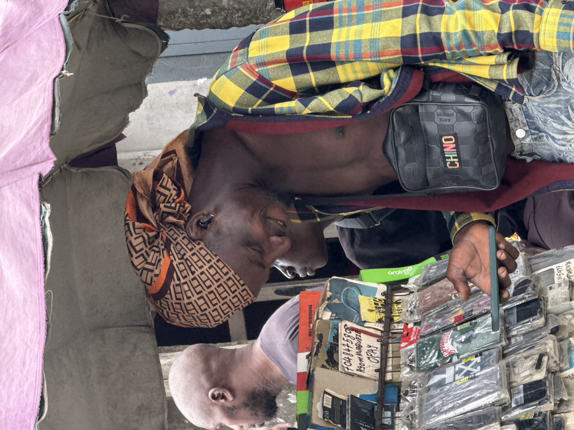 A medium shot shows two men at a market stall in Ado, Nigeria. The first man, who is dark-skinned, wears a printed headscarf and a yellow and blue plaid shirt with an unbuttoned front, a black Michael Kors CHINO bag, and holds a green phone in front of phone cases wrapped in plastic on a display rack. The second man, partially visible in the lower left, has a shaved head, dark beard, and wears a lavender shirt. The stall displays advertisements, including a handwritten sign with the phone number "7048475808" and the words "ASOM NWORBUEZE OPAY", and a green box with "oraimo" printed on it. A pink curtain or drape hangs on the left. The background is a mix of fabric and a concrete surface.