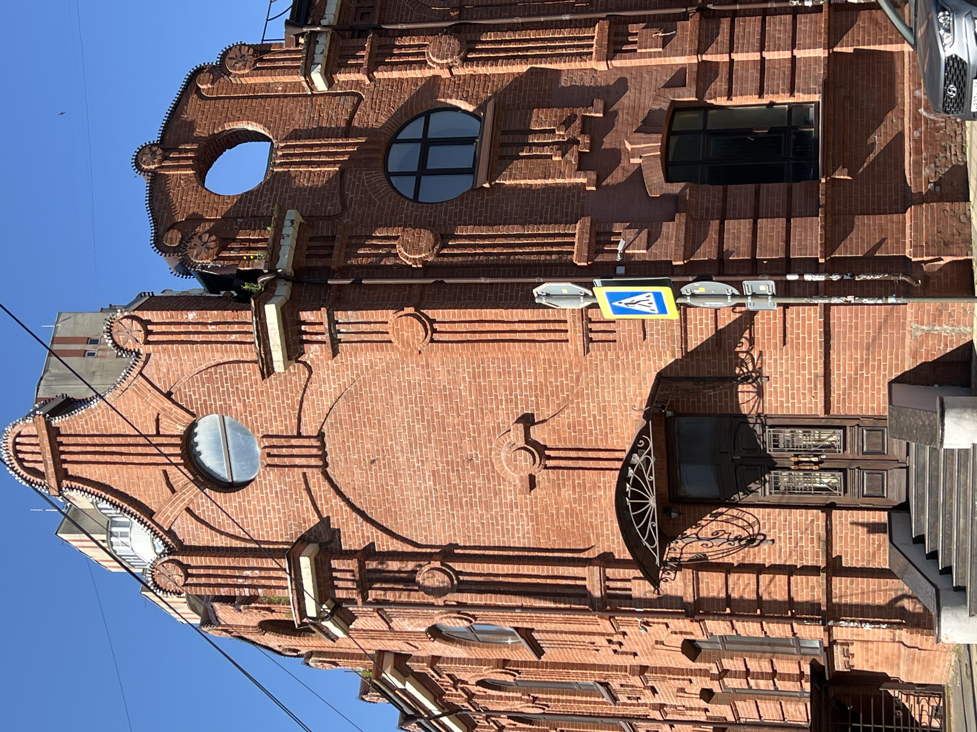 This image shows the exterior of a red brick building, likely a historical structure, located in Krasnodar, Russia. The building features ornate architectural details, including decorative brickwork, arched window frames, and a unique, large circular opening near the roofline on the right side. The entrance to the building is marked by a double wooden door with decorative metal grilles on the windows, accessible by a set of stairs. A metal awning with an intricate design is situated above the entrance. There are several windows of various shapes and sizes on the facade. A pedestrian crossing sign is visible on the sidewalk in front of the building. Overhead wires are strung across the bright blue sky. A car is partially visible in the lower right corner of the image. The sunlight casts strong shadows, indicating it is daytime.