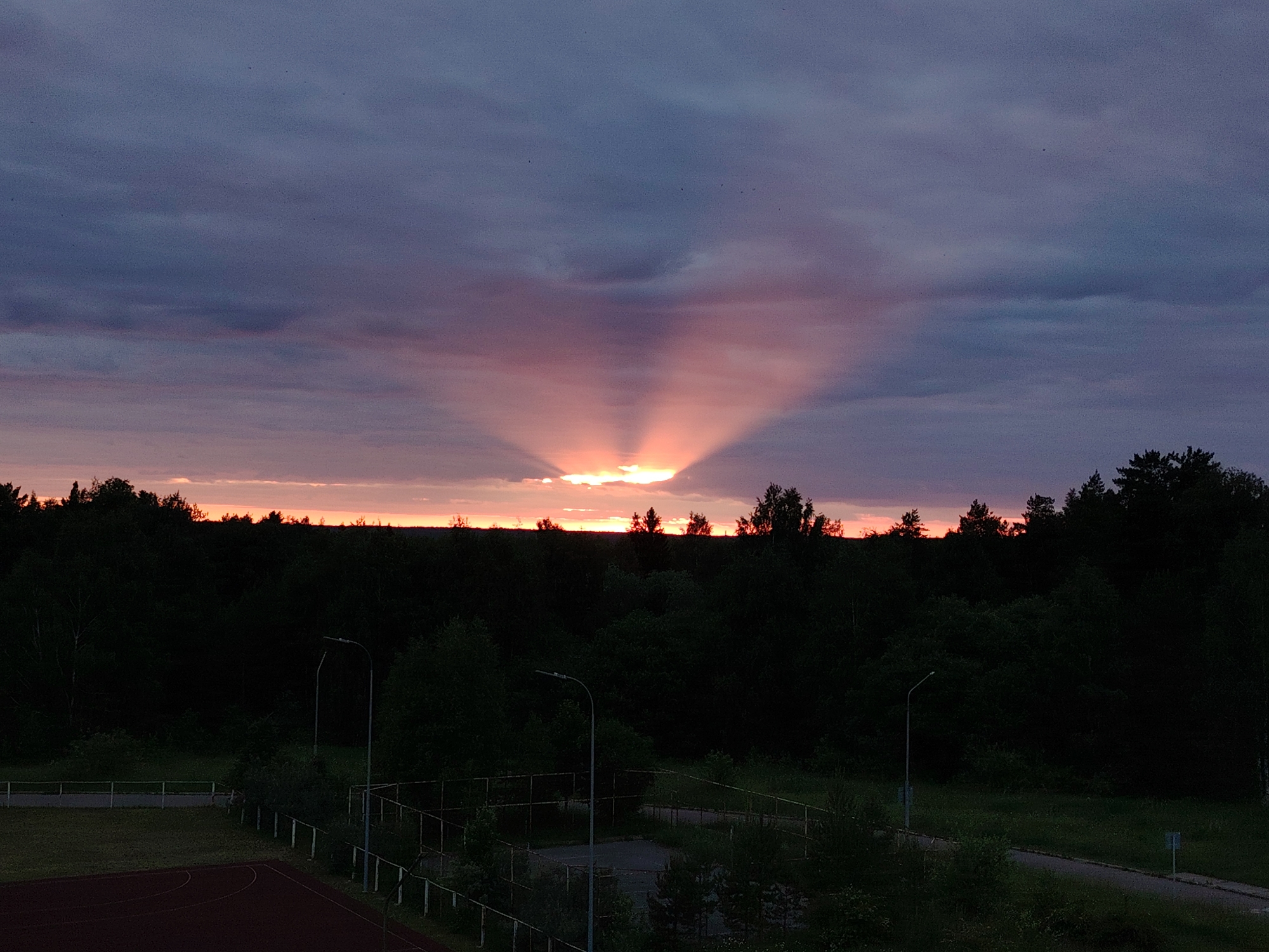 The image depicts a twilight scene in the city of Markovskiy, Russia. The sky is filled with dramatic crepuscular rays emanating from a low-lying sun, partially obscured by clouds. The colors in the sky range from deep purples and blues at the top to oranges and pinks closer to the horizon. Below the sky is a dense, dark silhouette of a forest. In the foreground, a sports field with a red track and green grass is visible, along with a fenced-in area that appears to be a small court. Two streetlights stand tall amidst the greenery and the structures of the sports facility. The overall impression is one of a serene, natural landscape at dusk, with hints of human infrastructure.