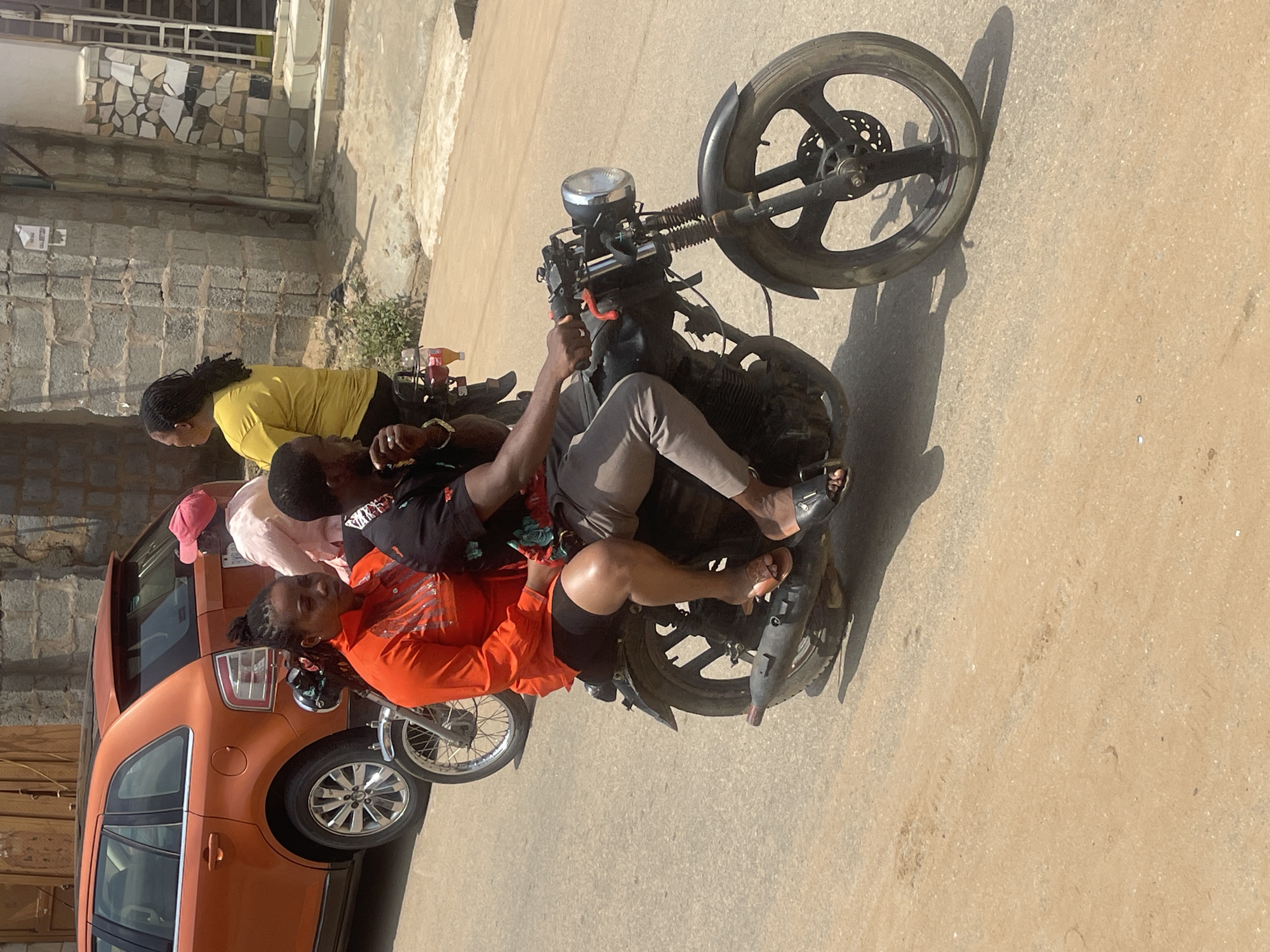 This image captures a vibrant scene in Abuja, Nigeria, likely during the daytime given the bright sunlight and harsh shadows. The primary focus is a group of people gathered around a motorcycle. One person, wearing an orange top and black shorts, is seated on the motorcycle, looking towards the camera with a relaxed expression. Another person, in a yellow top, stands behind the motorcycle, facing away from the camera. A third individual, in a pink cap, is partially visible behind them.
The motorcycle itself is dark and appears to be a standard model, with visible handlebars, a headlight, and spoked wheels. It is parked on a paved surface, likely a street or road, with a portion of an orange car visible on the left side of the frame. The car's silver rim is a notable detail. In the background, there are elements of urban architecture, including a stone wall and what looks like a building with a metal railing. A small patch of greenery is also visible.
The overall atmosphere seems casual and social, with the individuals appearing to be either preparing to ride, taking a break, or simply conversing. The lighting suggests a sunny day, and the warmth of the sunlight is palpable. The presence of the car and the urban background firmly place the scene in a populated area. There is no visible text in the image.