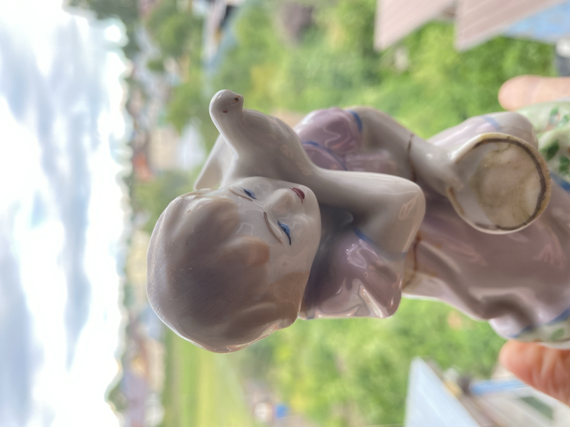 A close-up, side-angle shot shows a porcelain figurine of two figures, possibly children, embracing. The figurine is predominantly white with hints of pink and blue. The face of one figure, closer to the camera, is detailed with closed blue eyes and pink cheeks. The figures are positioned in a way that suggests affection or rest. The background is softly blurred, showing a landscape with green foliage and hints of buildings, suggesting an outdoor setting during daytime. The lighting appears natural and bright, indicating it might be midday. The image is captured with a shallow depth of field, focusing on the figurine while blurring the surroundings. The context provided indicates the location is Zelenodol'sk, Russia. There is no visible text in the image.