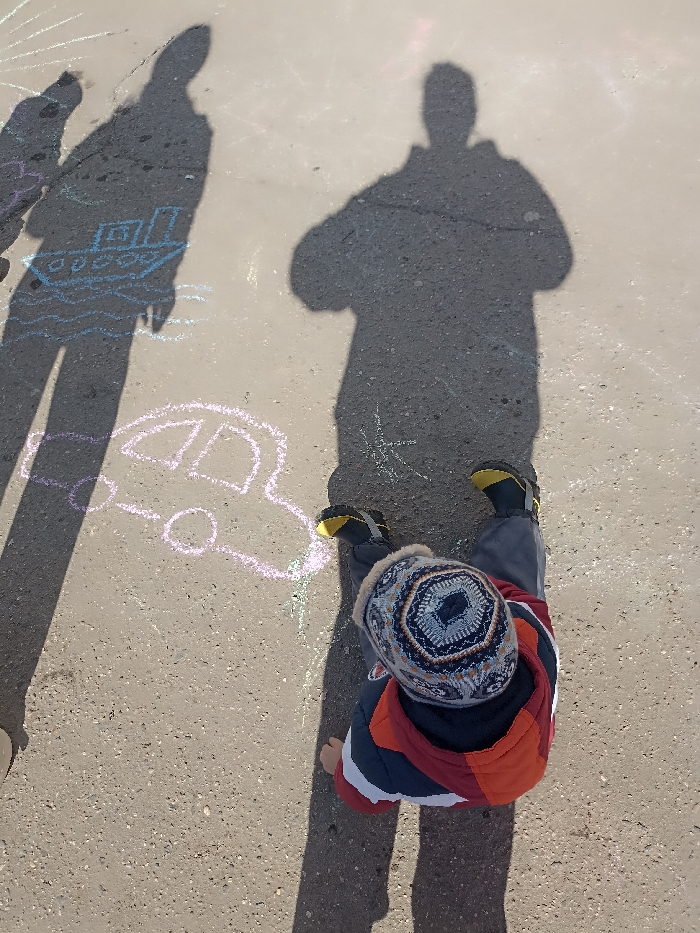The scene is an outdoor paved area in Serpukhov, Russia, under strong sunlight casting distinct shadows. A young child, viewed from above, kneels or squats on the light-grey asphalt surface. The child wears a multi-colored knitted hat with geometric patterns, a color-block jacket (red, navy, white), grey pants, and dark shoes with prominent yellow soles. Two elongated human shadows are cast onto the ground, indicating the presence of at least two adults. One slender shadow is on the left side of the frame, and a broader shadow is on the right, both positioned behind and to the side of the child. The asphalt surface features multiple chalk drawings; a light blue drawing of a boat on waves is visible near the left shadow, and a light pink drawing of a car is located on the ground between the child's feet and the left shadow. Other faint, colored chalk marks are present across the surface.