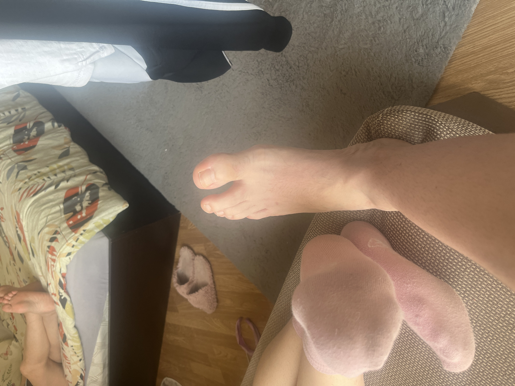 This image captures an indoor scene in Burgas, Bulgaria. In the foreground, a person's legs are shown, with the right foot bare, exhibiting natural toenails and fine hair on the skin. The left foot is covered by a light pink ankle sock, with an additional light pink sock draped over it, both resting on a textured, light brown upholstered surface, likely a sofa or armchair.
In the background, a bed with a dark brown frame is visible. On the bed, another person's bare feet are positioned, resting on a light blue-grey sheet under a duvet adorned with a patterned cover featuring light yellow, orange, black, and white abstract floral or leafy motifs. The floor is composed of light-colored wooden planks, partially overlaid by a light grey shaggy rug.
Located on the wooden floor are a pair of light pink, fluffy slippers and a small, circular, light purple hair tie. To the far right, a white t-shirt and a black jacket with a prominent white stripe are hanging or draped. The scene is illuminated by natural daylight, suggesting a daytime setting.