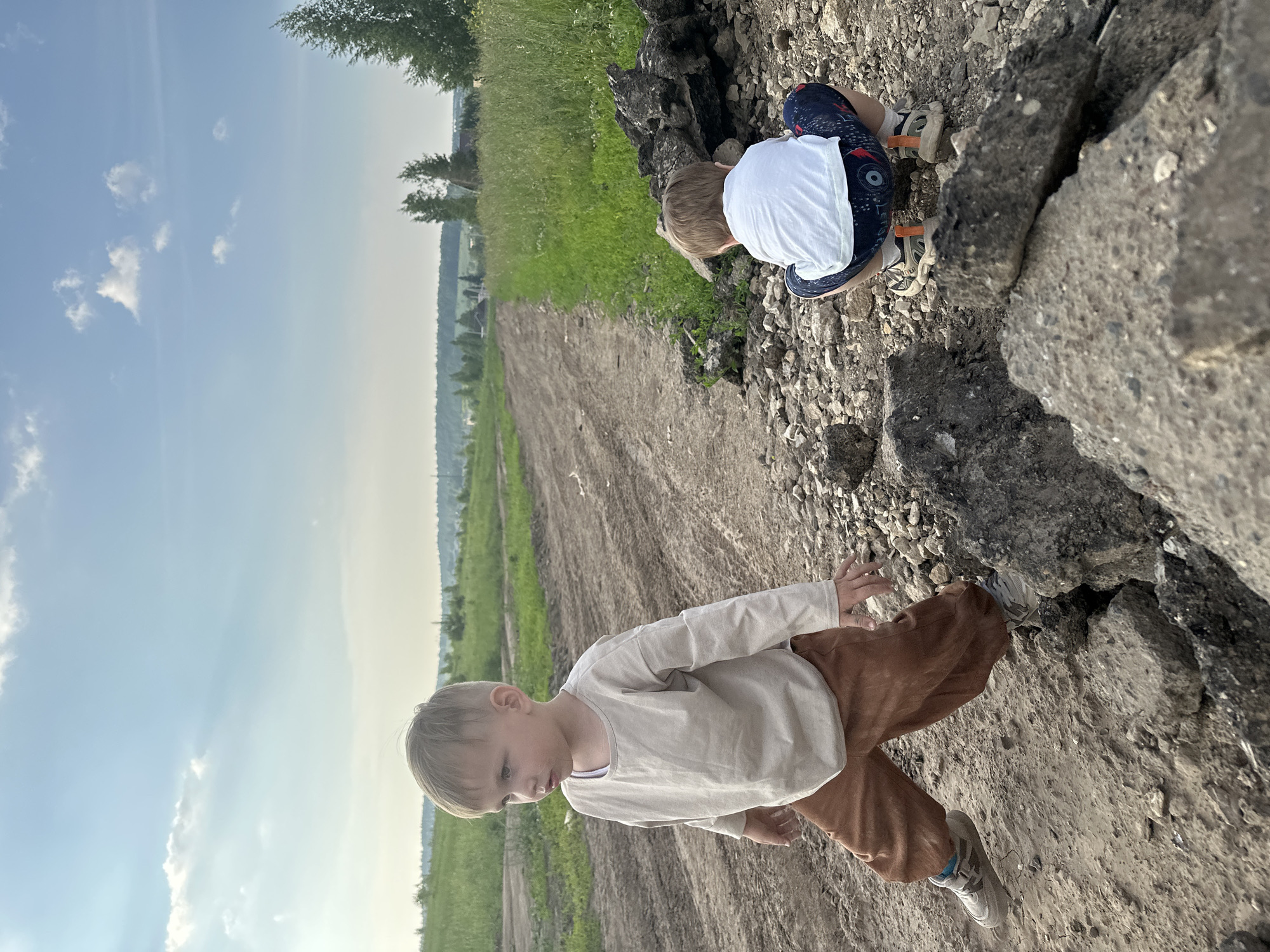 Two young boys are in a rural outdoor environment, likely in the vicinity of Estachi, Russia. The scene depicts a dirt road that stretches into the distance, flanked by grassy fields and scattered trees. In the foreground, a dirt pile composed of rocks and what appears to be broken asphalt is visible.
The boy on the left is facing towards the right of the frame, mid-stride. He has light blonde hair and is wearing a loose-fitting, long-sleeved beige shirt and brown pants. His right hand is extended outwards.
The boy on the right is positioned in a crouched stance, facing away from the camera. He has blonde hair and is wearing a white t-shirt and patterned shorts. His feet are clad in sneakers with orange accents.
The sky above is partly cloudy with a clear blue expanse. The overall lighting suggests it is daytime, possibly late afternoon or early evening given the warm tones. The environment appears undeveloped and somewhat rugged.