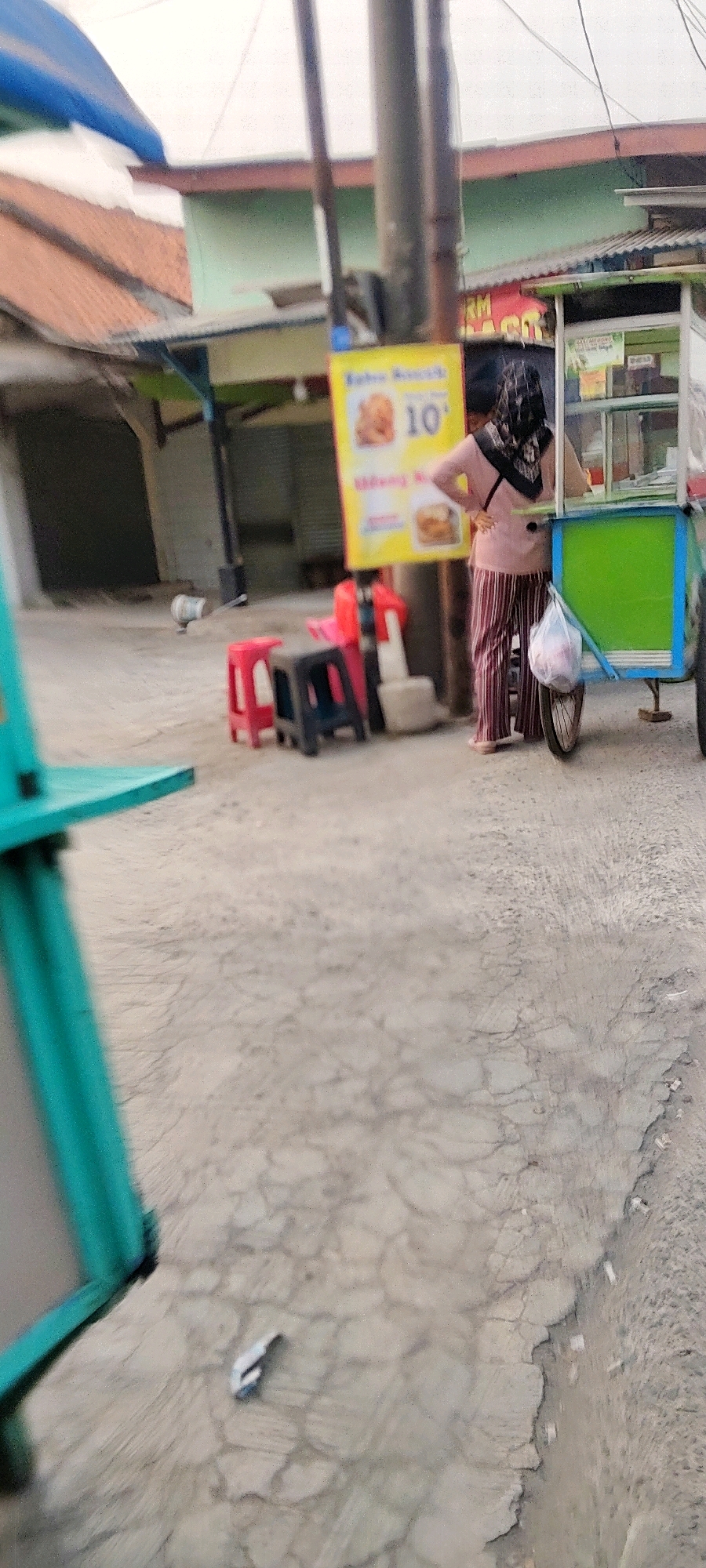 In Mekarmukti, Indonesia, a street scene depicts a person standing next to a food cart. The person, wearing a headscarf, a pink top, and striped pants, stands with their hands on their hips. A plastic bag hangs from the side of the food cart, which is green and blue. Several red and black stools are arranged on the cracked concrete ground, with a yellow sign displaying "10" in the background. In the distance, buildings with orange and green roofs are visible, along with utility poles. A small white object, possibly trash, lies on the ground near a white pole. A teal-colored object is in the foreground on the left. The overall impression is a casual street scene with food vending activity.