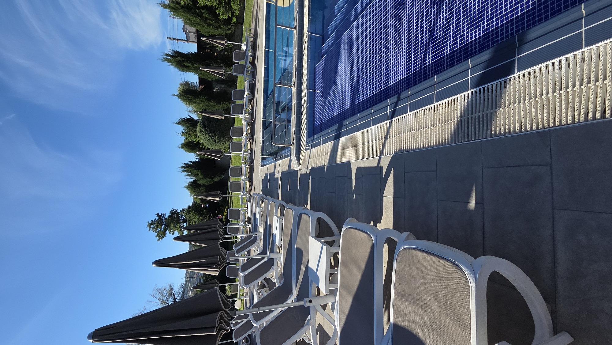 The image depicts an outdoor swimming pool area on a clear day, likely in a resort or hotel. A row of closed, dark gray umbrellas stands behind a series of empty, white-framed lounge chairs with brown cushions. The lounge chairs are arranged on a stone patio. To the right, a portion of a swimming pool is visible, with bright blue tiles lining the bottom and sides. The pool is empty. In the background, a line of evergreen trees separates the patio area from a residential or wooded area, with a few buildings visible in the distance. The sky is a vibrant blue with some wispy white clouds. The sun is casting long shadows from the umbrellas and lounge chairs, indicating it is either morning or late afternoon.