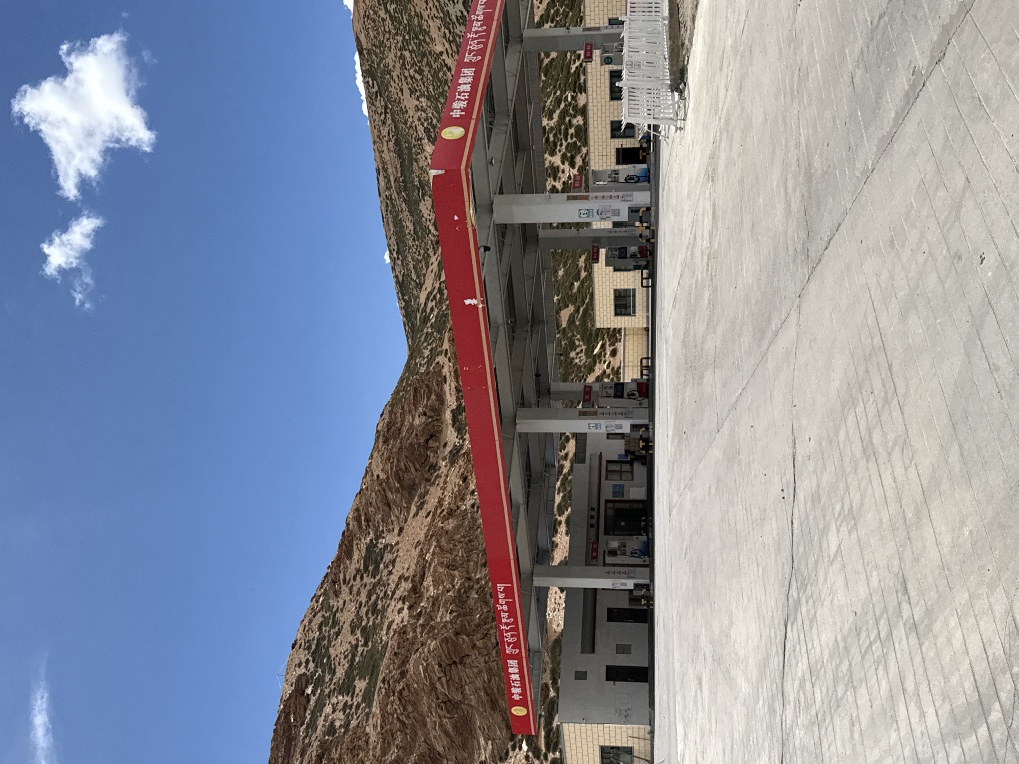 The image shows a Sinopec gas station in Kela Township, China. The station is painted red and white and sits next to a rocky mountain under a blue sky with some clouds. A concrete pad stretches out in front of the gas station.