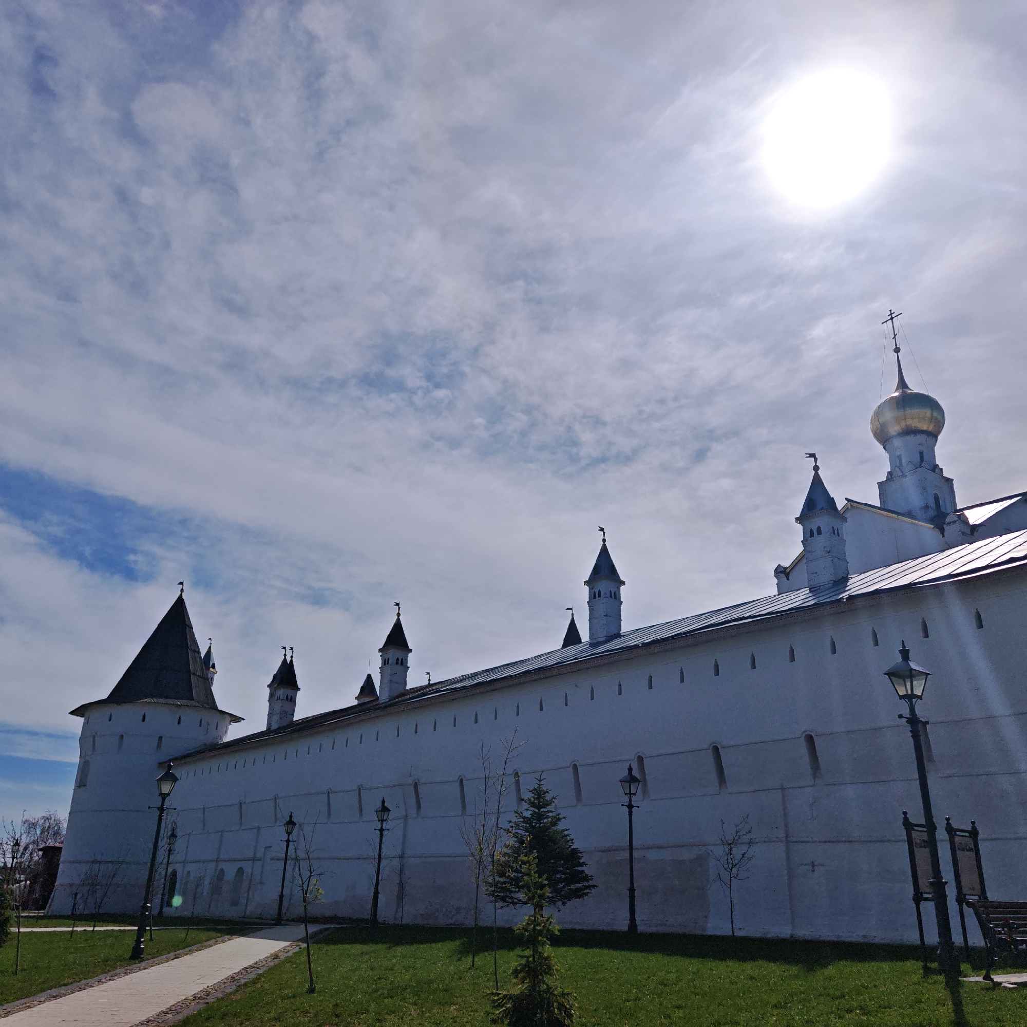 The image shows a daytime view of the Spaso-Yakovlevsky Monastery in Rostov, Russia. The monastery's white walls with turrets are visible, along with a golden-domed church tower. The sky is partly cloudy with the sun high above. In the foreground, there's a grassy area with small trees and lampposts along a paved pathway. A bench and what appears to be a sign are present to the right of the frame.